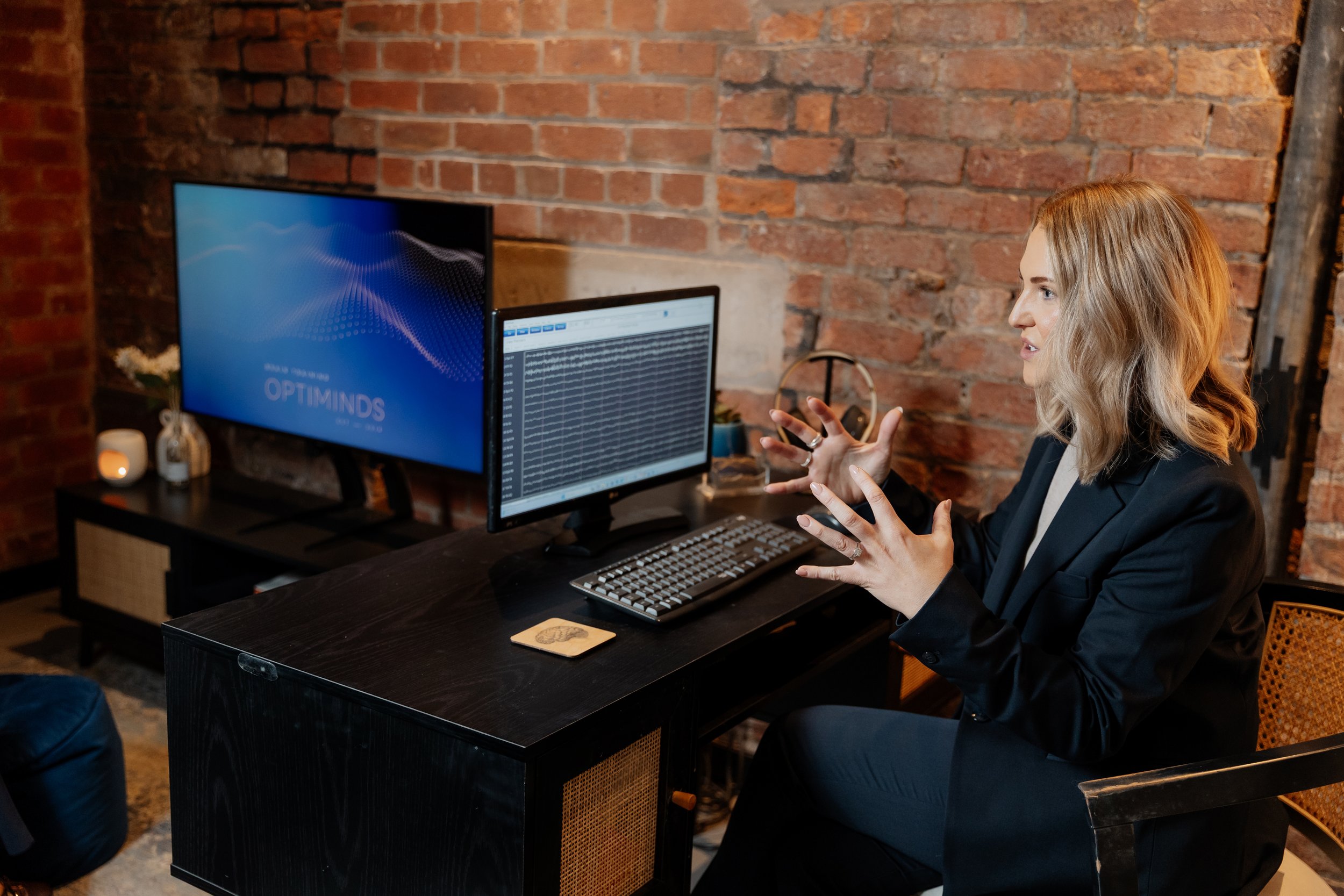 Woman with blonde hair in a black blazer talking in front of dual computer monitors in a modern office with exposed brick wall.