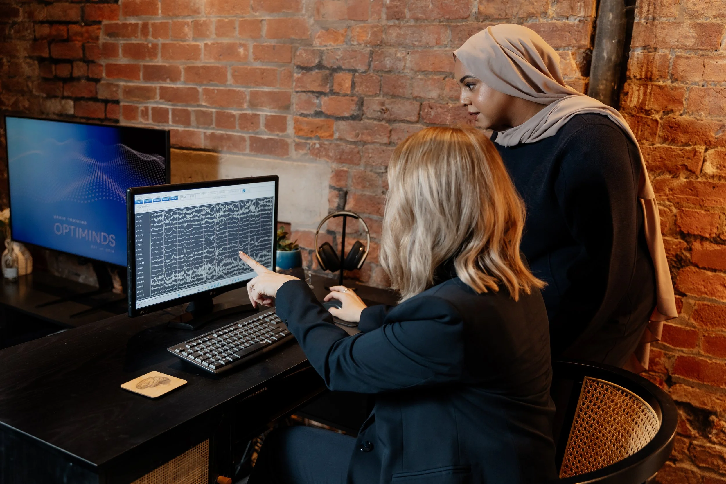 Two women working together at a desk with computer screens showing brainwave data. One woman in a hijab points at the monitor while the other woman observes. The workspace has a brick wall background.