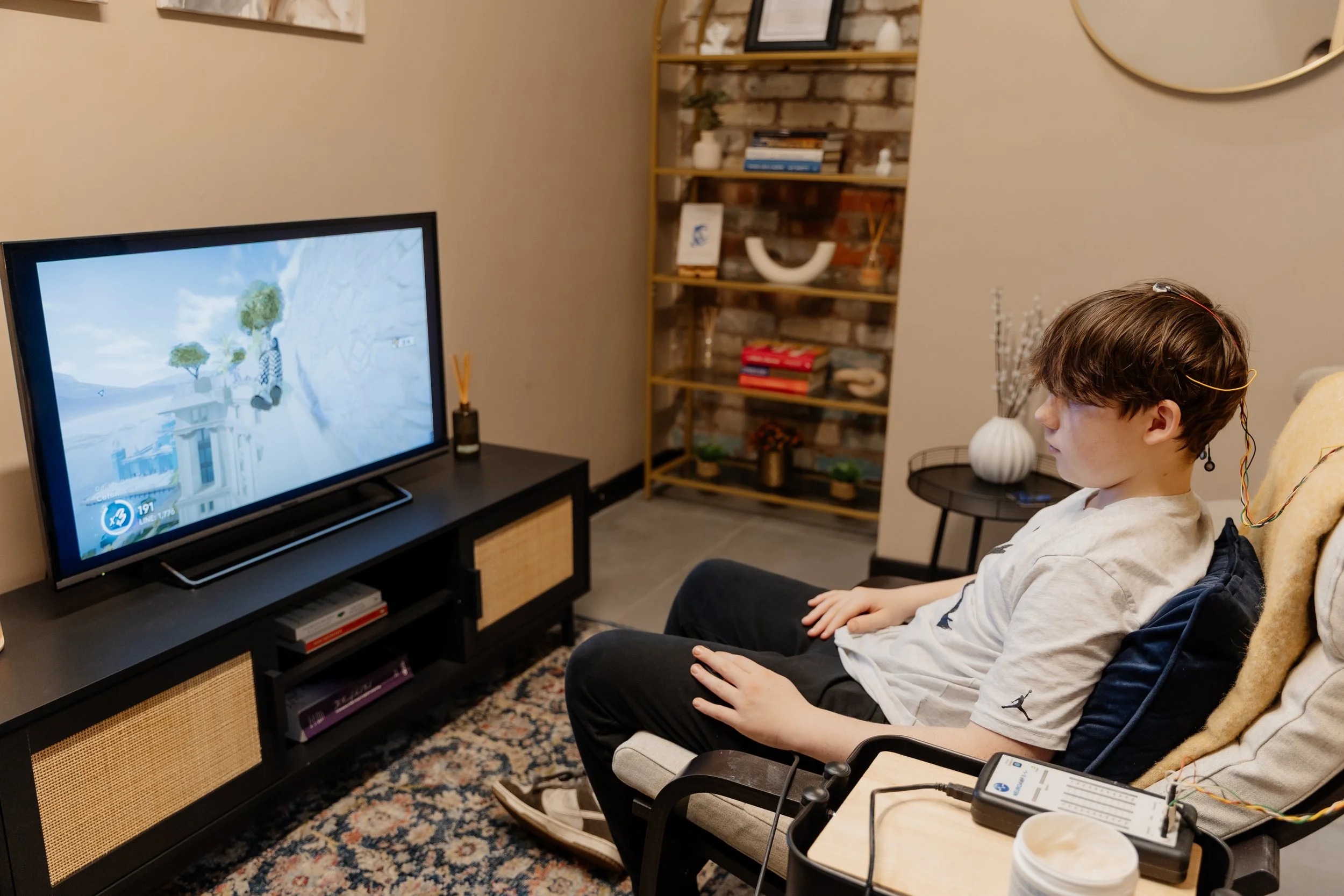 A boy with brown hair sitting in a chair, playing a video game on a TV. The room has a mantle with bookshelf, books, decorative items, a mirror, and a vase. There is a patterned rug on the floor.