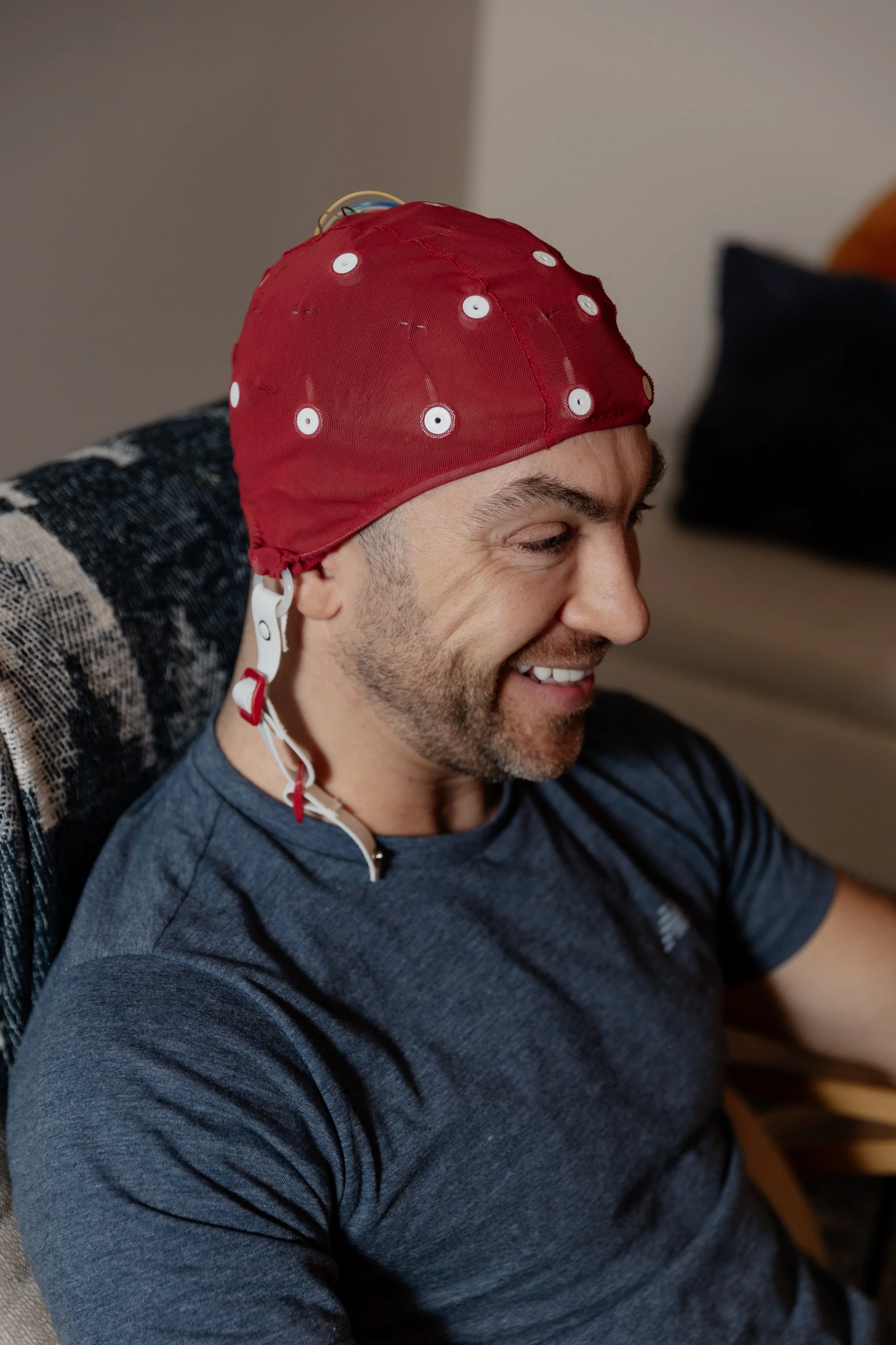 A man wearing a red QEEG cap with sensors, smiling and sitting on a chair.