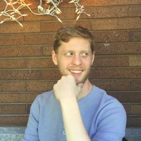 A young man with light brown hair and a beard smiling, wearing a blue shirt, sitting outside against a brick wall with some leafless branches above.