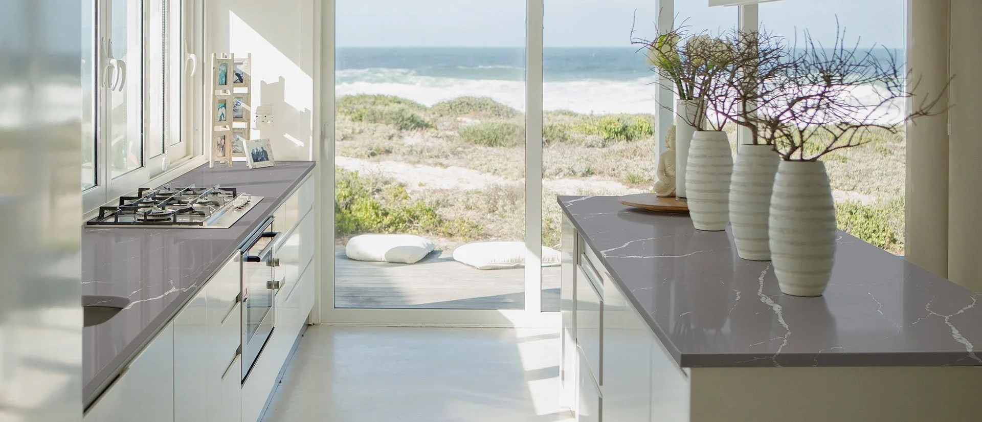Modern kitchen with marble countertops, potted plants, and a view of the beach through sliding glass doors.