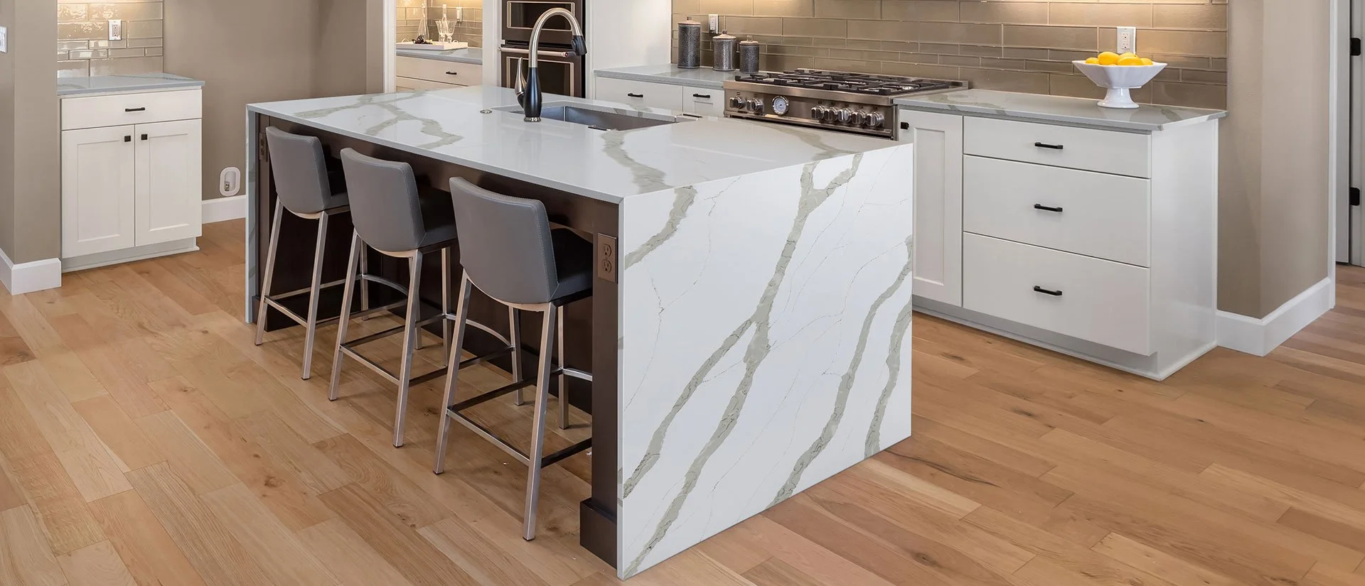 Modern kitchen with white marble island featuring grey and gold veining, gray barstools with metal legs, beige walls, light wood flooring, and white cabinetry.