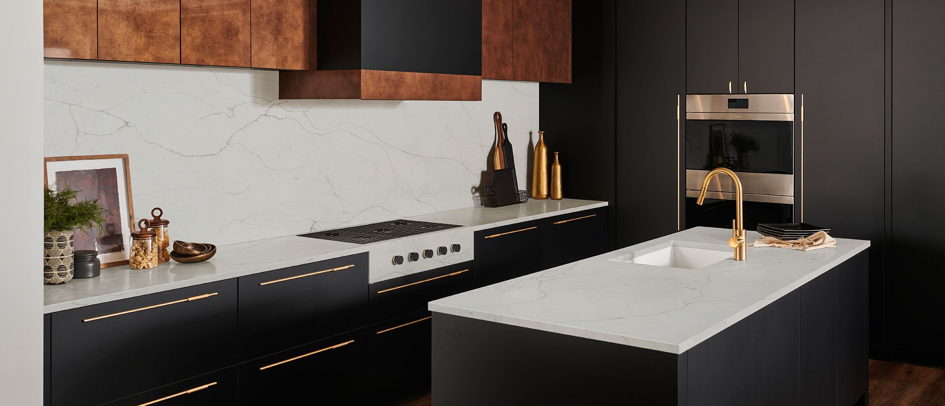 Modern kitchen with black and copper cabinetry, white marble countertops, and a marble backsplash. Features a built-in oven, a gold faucet, and decorative items like gold vases, a plant, framed photo, and kitchen tools.