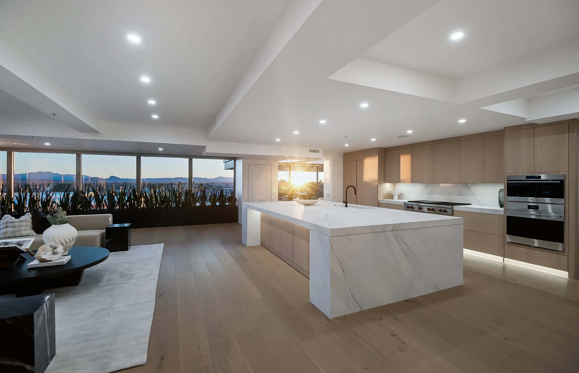 Modern minimalist kitchen with white cabinetry, a marble island, and integrated appliances. Natural light enters through large windows and a skylight. The room features light wood accents and a wooden dining table in Scottsdale.