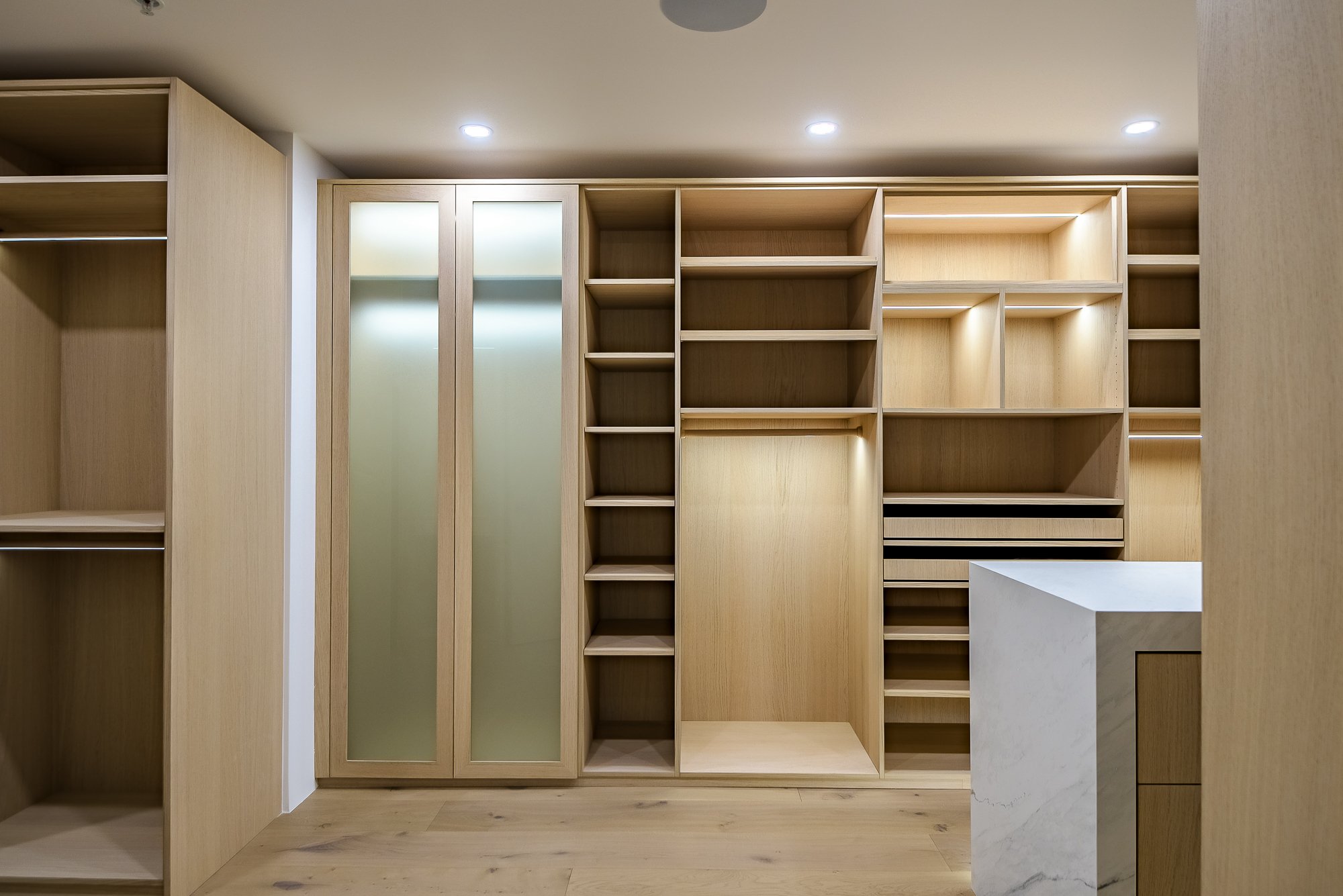 Modern walk-in closet with neutral tones, open shelving with folded clothes, hanging garments, a sliding glass door leading to outdoor desert landscape, and a countertop with a potted plant and hanging pendant lights.