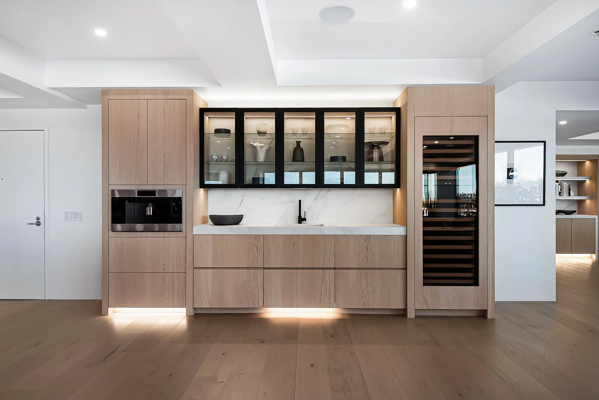 White oak dry bar with integrated wine fridge and beverage cooler, featuring glass upper cabinets and warm under-cabinet lighting