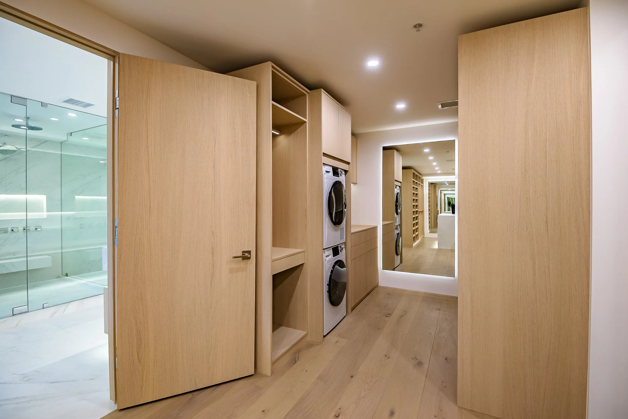 Modern walk-in closet with neutral tones, open shelving with folded clothes, hanging garments, a sliding glass door leading to outdoor desert landscape, and a countertop with a potted plant and hanging pendant lights.