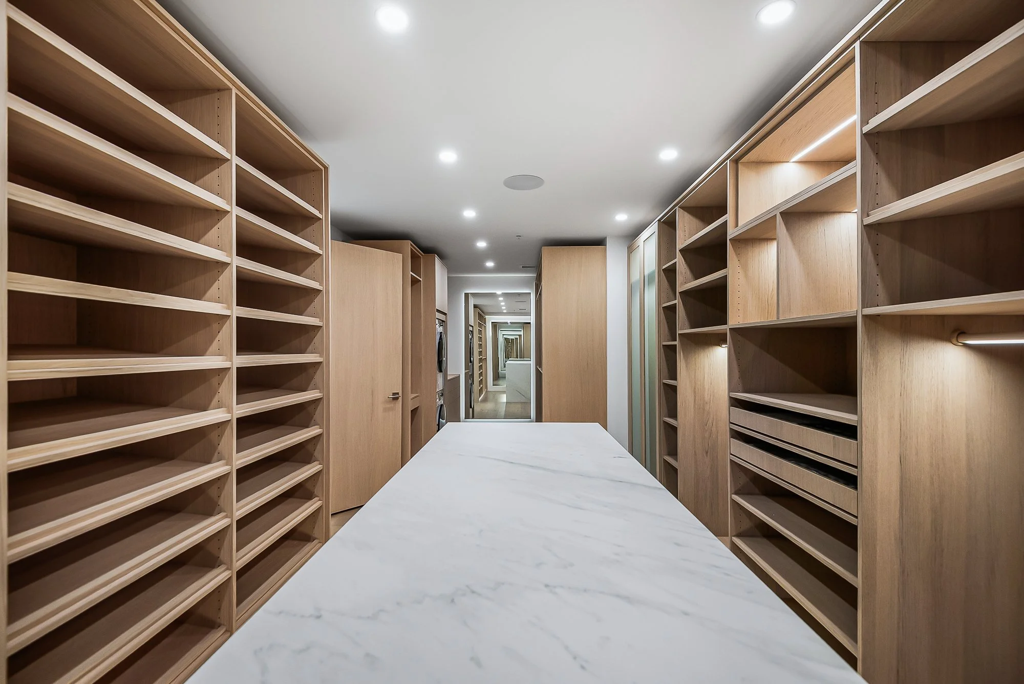 Modern walk-in closet with neutral tones, open shelving with folded clothes, hanging garments, a sliding glass door leading to outdoor desert landscape, and a countertop with a potted plant and hanging pendant lights.