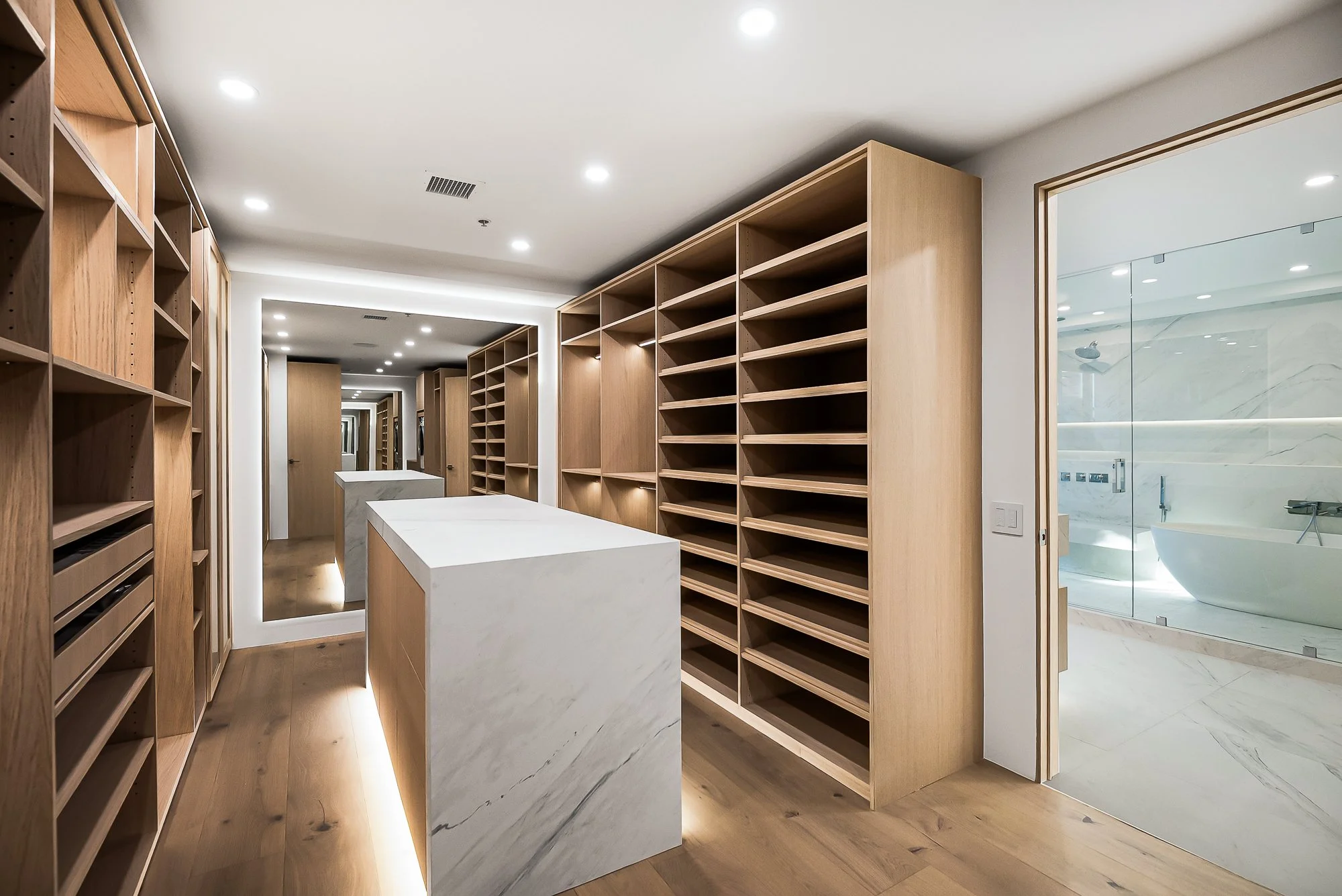 Modern walk-in closet with neutral tones, open shelving with folded clothes, hanging garments, a sliding glass door leading to outdoor desert landscape, and a countertop with a potted plant and hanging pendant lights.