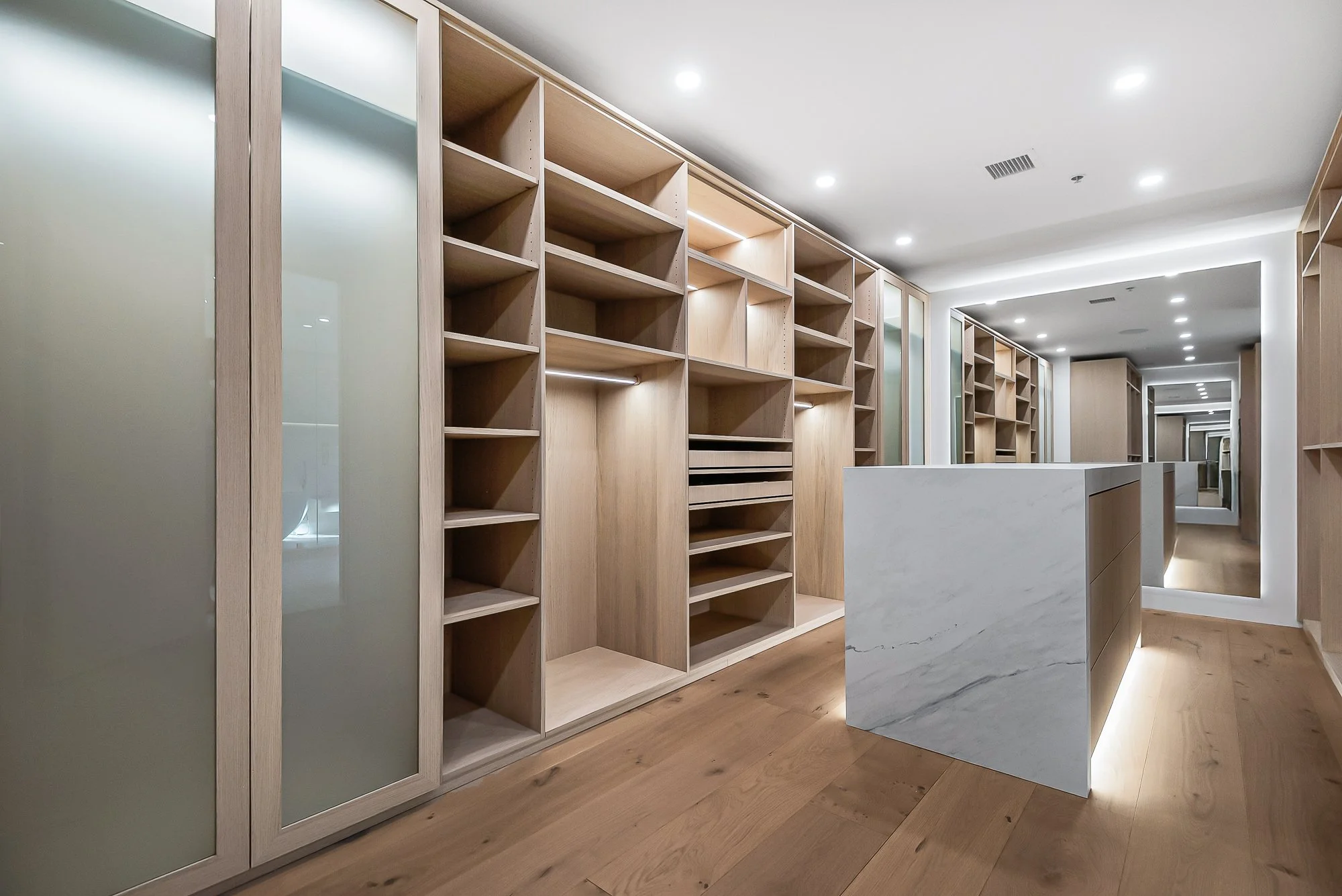 Modern walk-in closet with neutral tones, open shelving with folded clothes, hanging garments, a sliding glass door leading to outdoor desert landscape, and a countertop with a potted plant and hanging pendant lights.