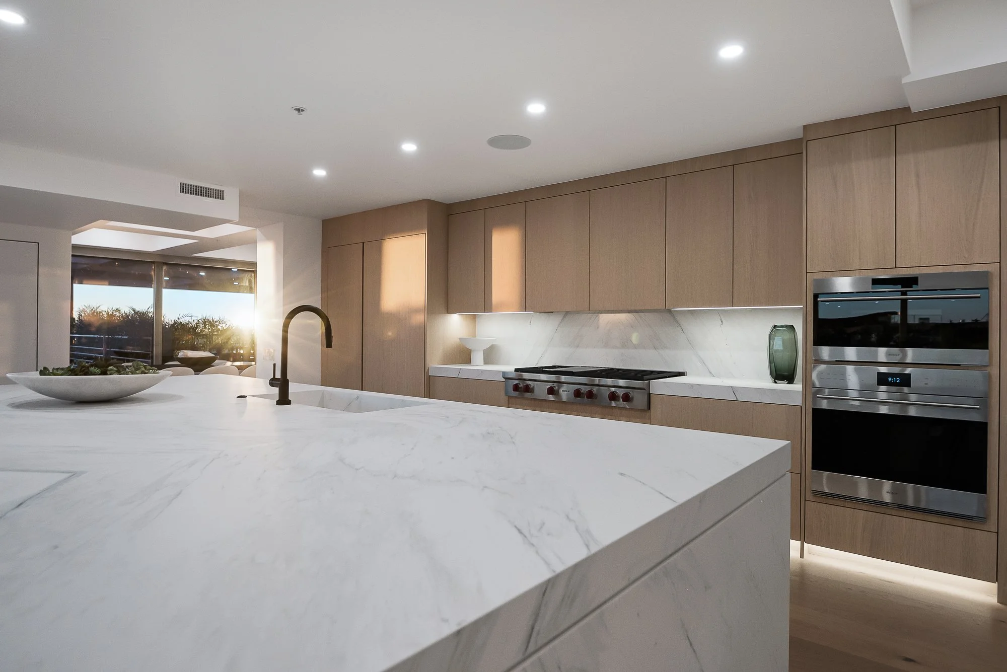 Modern kitchen with minimalistic design, featuring a marble island, light wood cabinetry, built-in ovens, and large windows.
