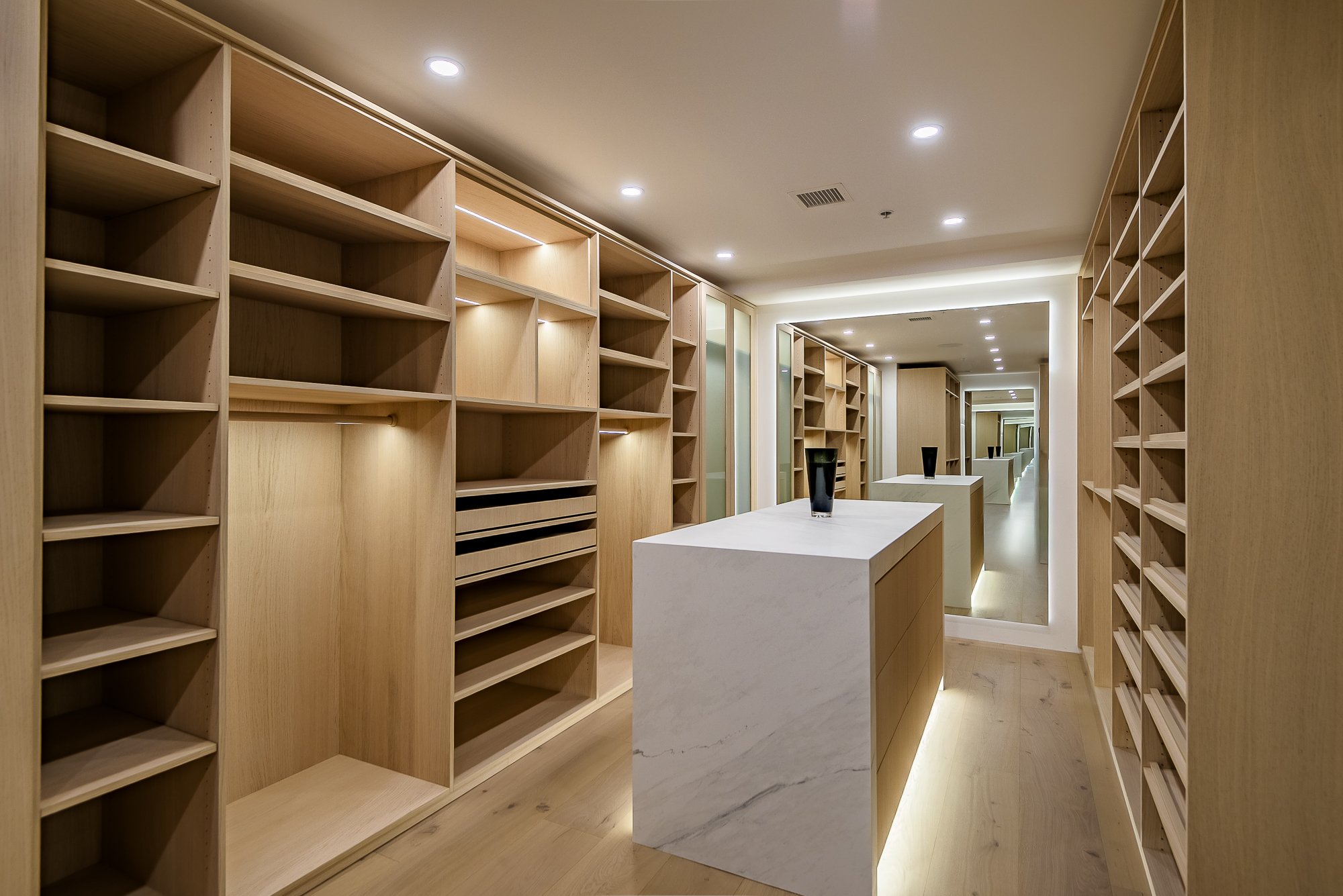 Modern walk-in closet with open shelves, hanging space for jackets, neatly folded towels, woven basket, and two cushioned ottomans facing large sliding glass doors leading to desert landscape.
