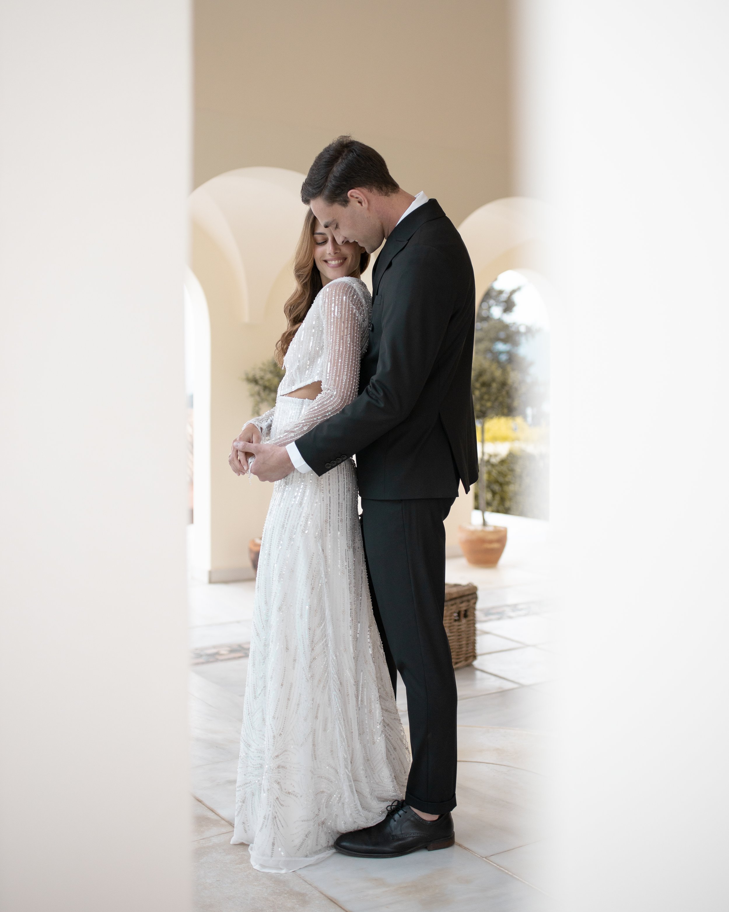 Bride and groom embracing indoors, elegant white dress, black suit, bright setting, romantic scene.