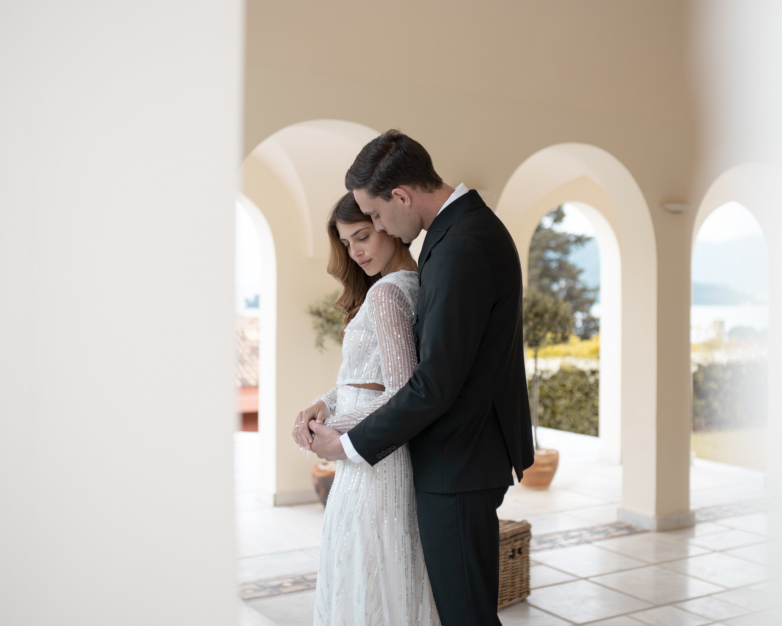 Bride and groom embracing in an elegant outdoor setting with arches and scenic views.