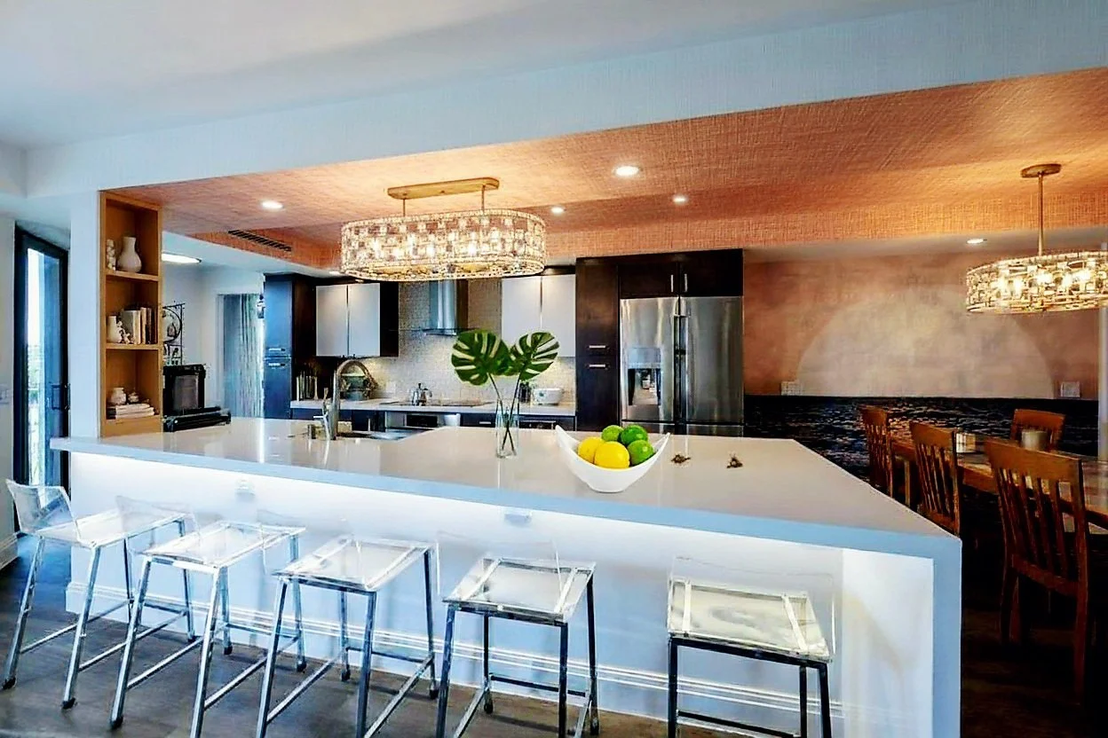Modern kitchen with white Quartz island countertop, stainless steel refrigerator, burnished brass metal backsplash, dark wood cabinets with wood and white frosted glass doors, and a decorative gilded and crystal chandelier overhead.