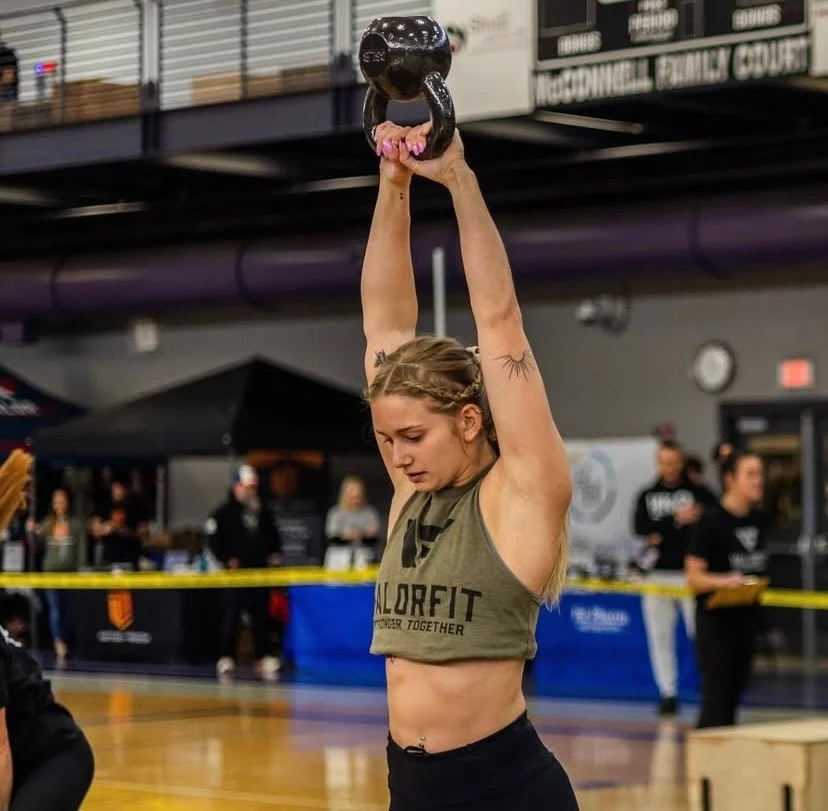 A woman lifting a kettlebell overhead in a gym setting, wearing a ValorFit tank top.