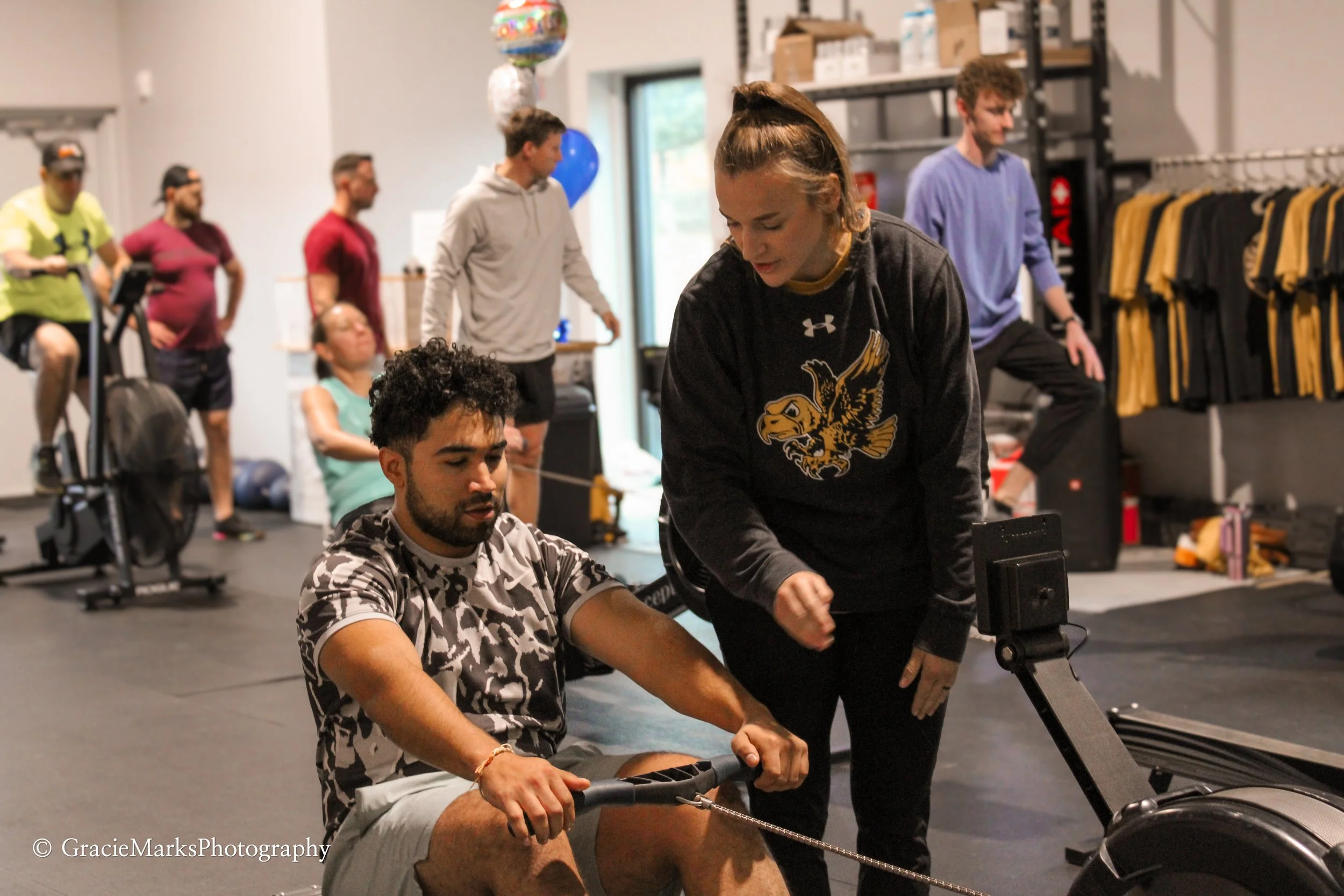 People working out in a gym with rowing and cycling machines; an instructor assisting
