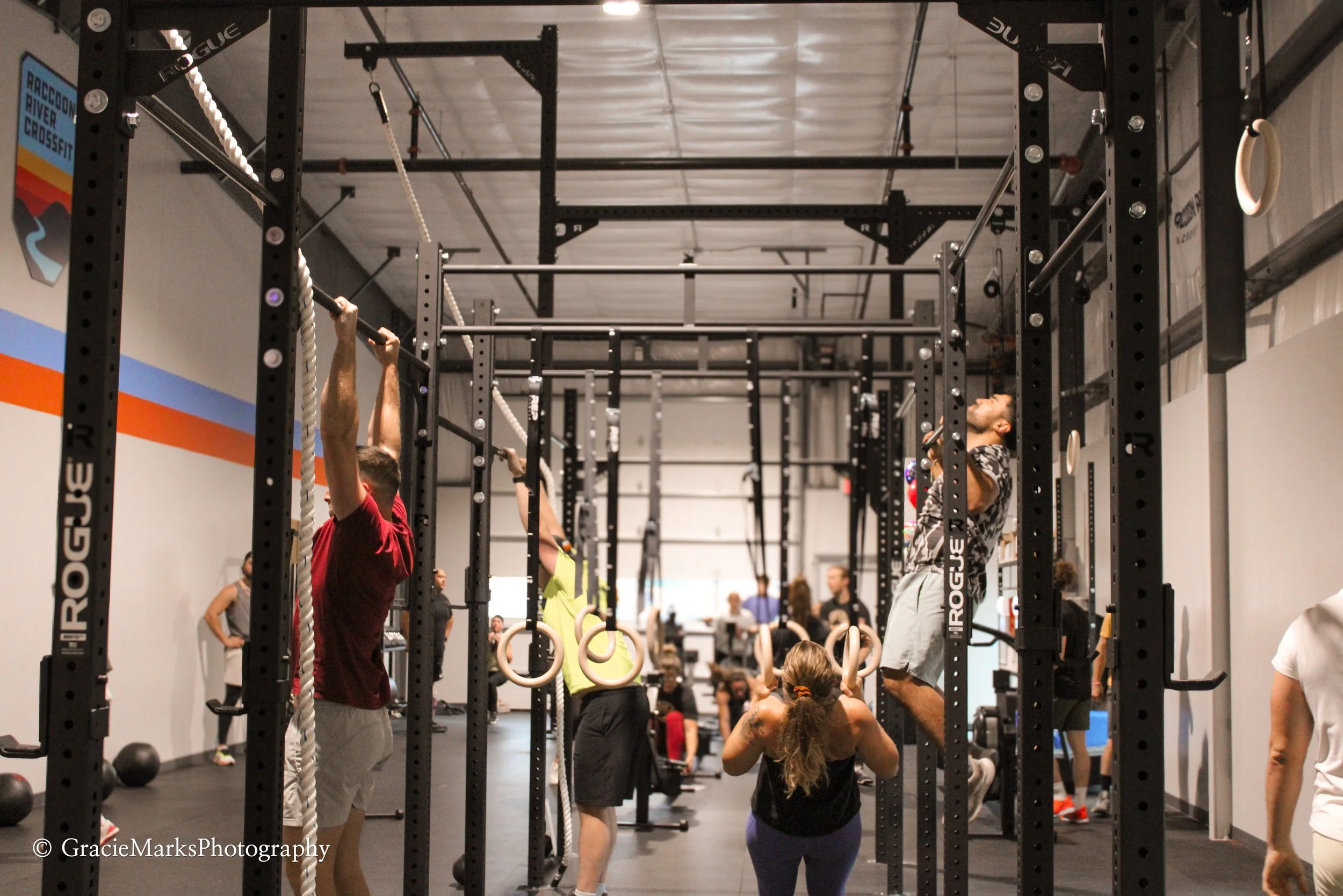 People exercising in a CrossFit gym, using pull-up bars and gymnastic rings, with Raccoon River CrossFit signage visible.