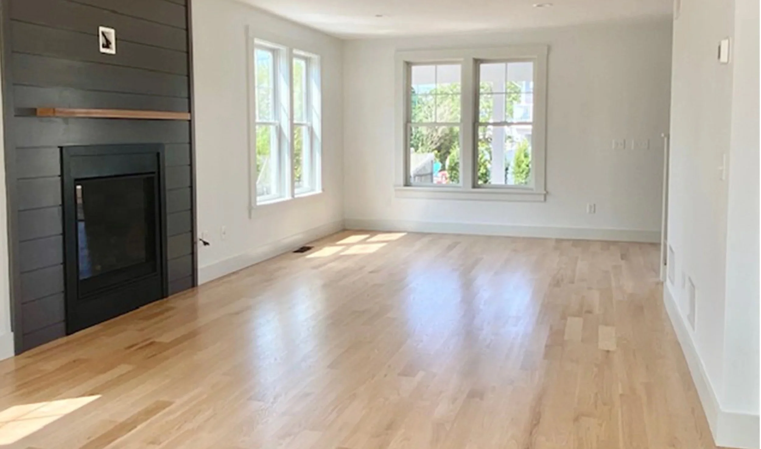Empty living room with large windows, hardwood floors, white walls, and a black fireplace with a wooden mantel.