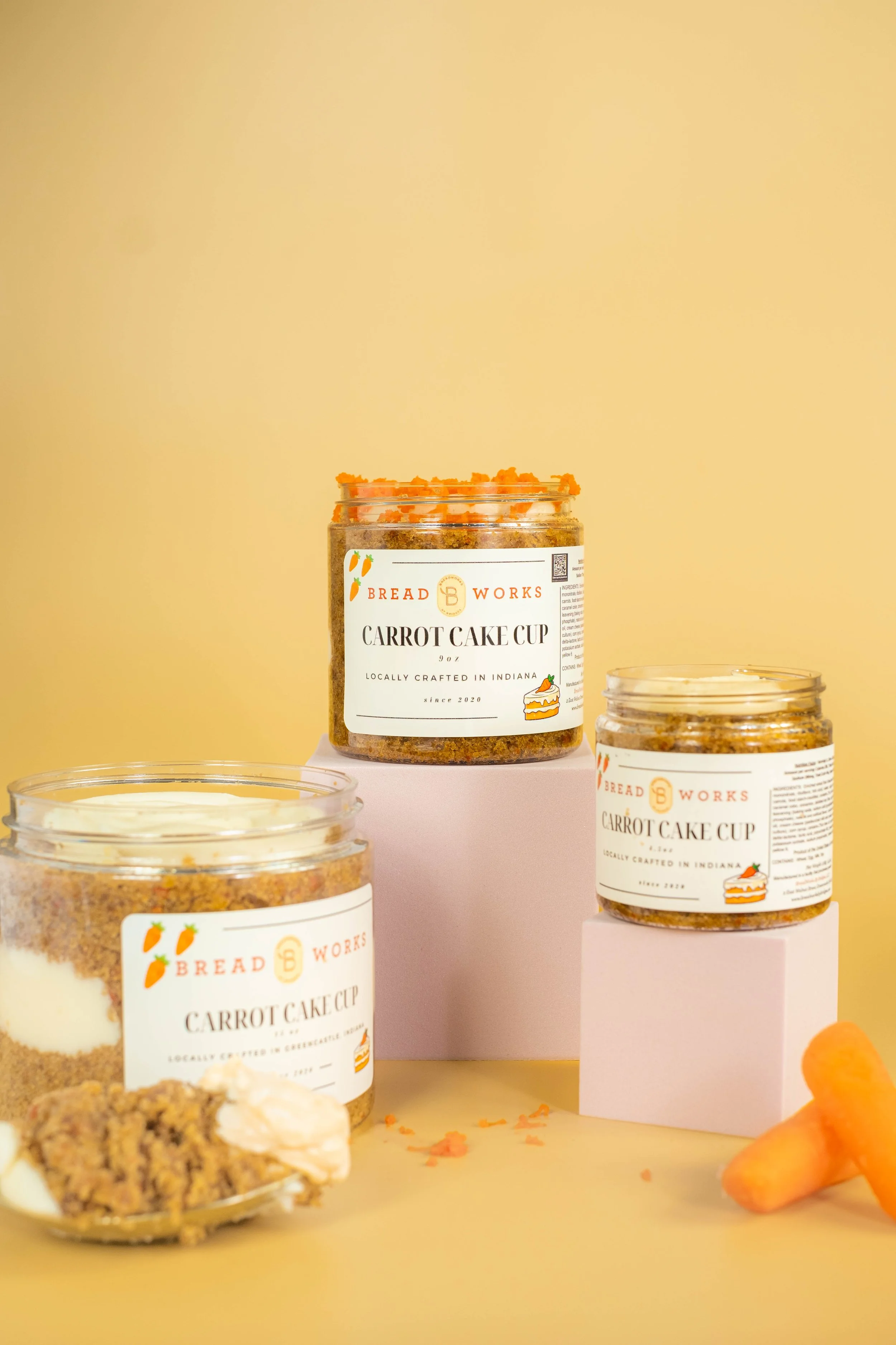 Three jars of carrot cake mix from Bread Works on a pink and yellow background, with some scattered carrot shavings and a baked carrot cake in the foreground.