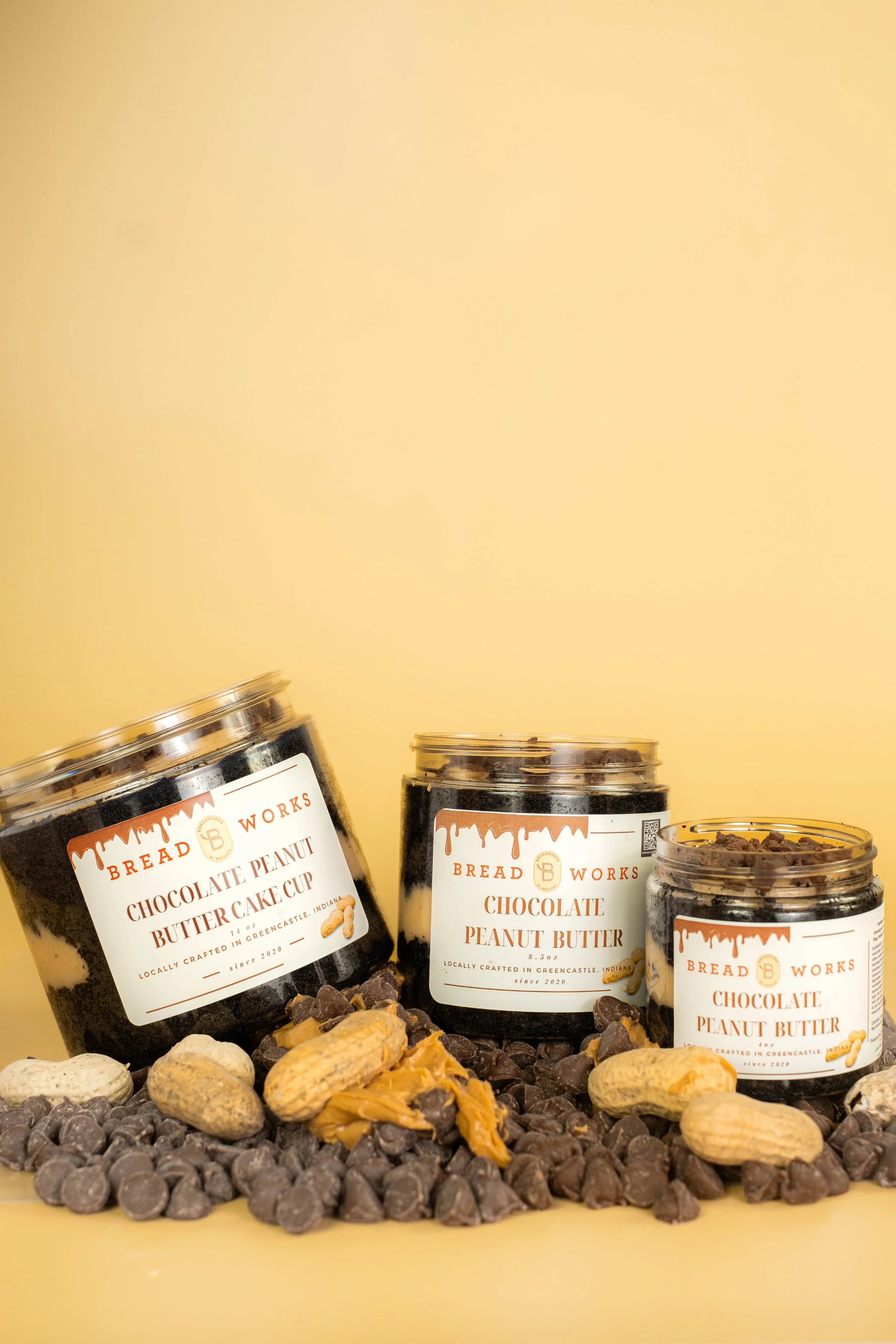 Three jars of chocolate peanut butter from Bread Works are arranged on a surface with assorted rocks and pebbles. The jars have white labels with orange text and a logo, indicating they are handcrafted in Greencastle, Indiana.