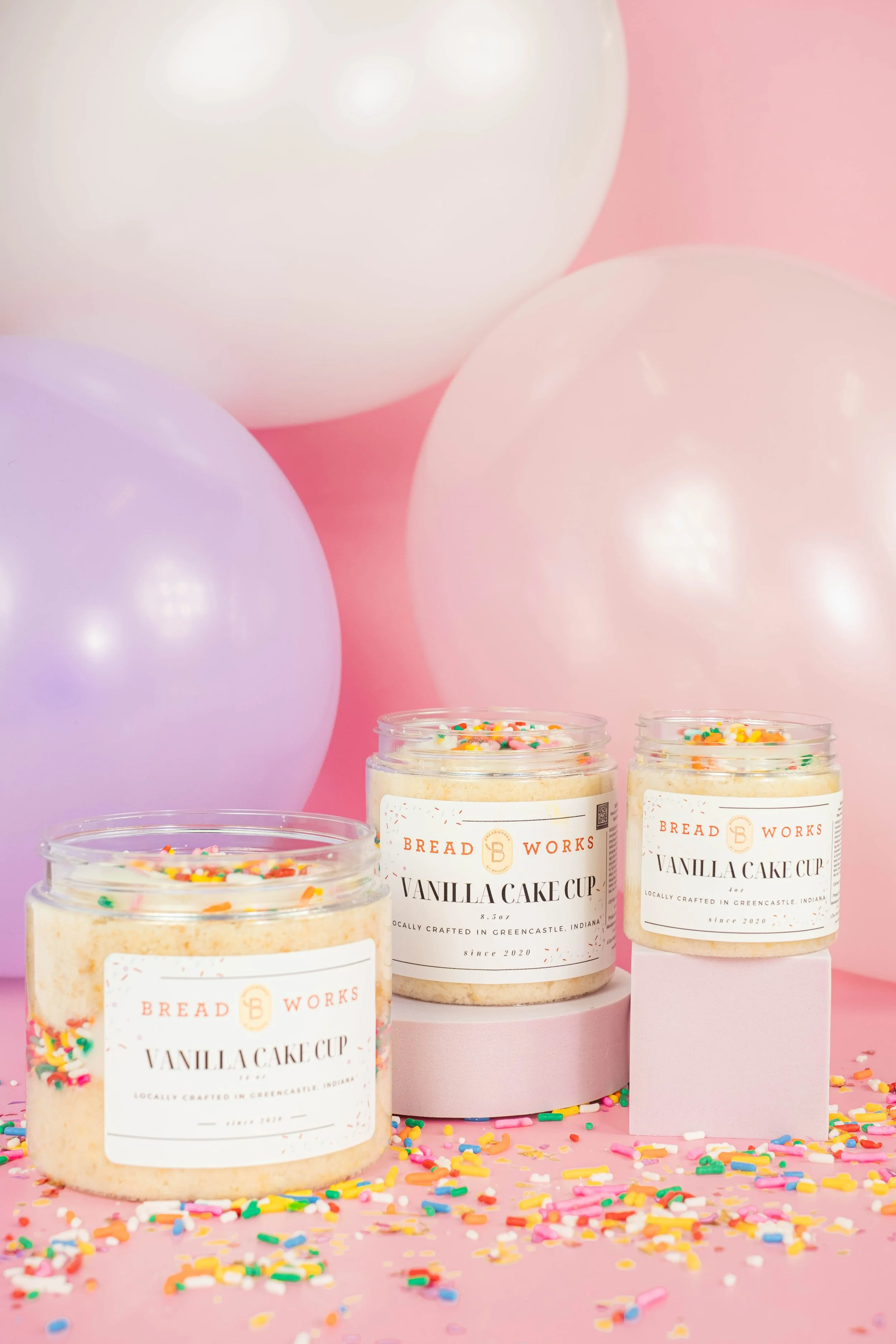 Three jars of vanilla cake cup mix with colorful sprinkles on a pink background, surrounded by scattered sprinkles and pastel-colored balloons.