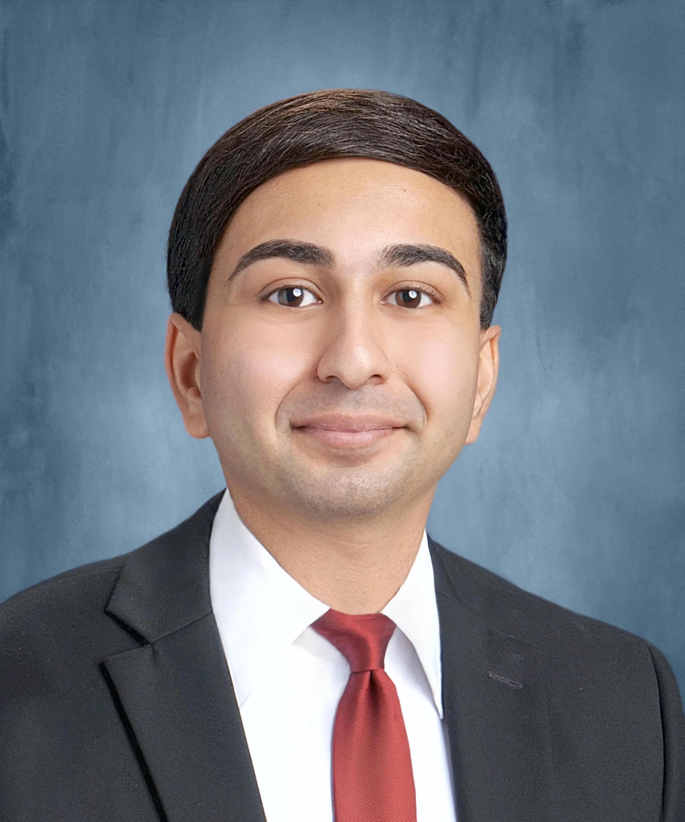 Professional portrait of a man with short dark hair, wearing a black suit and red tie, smiling with a blue background.