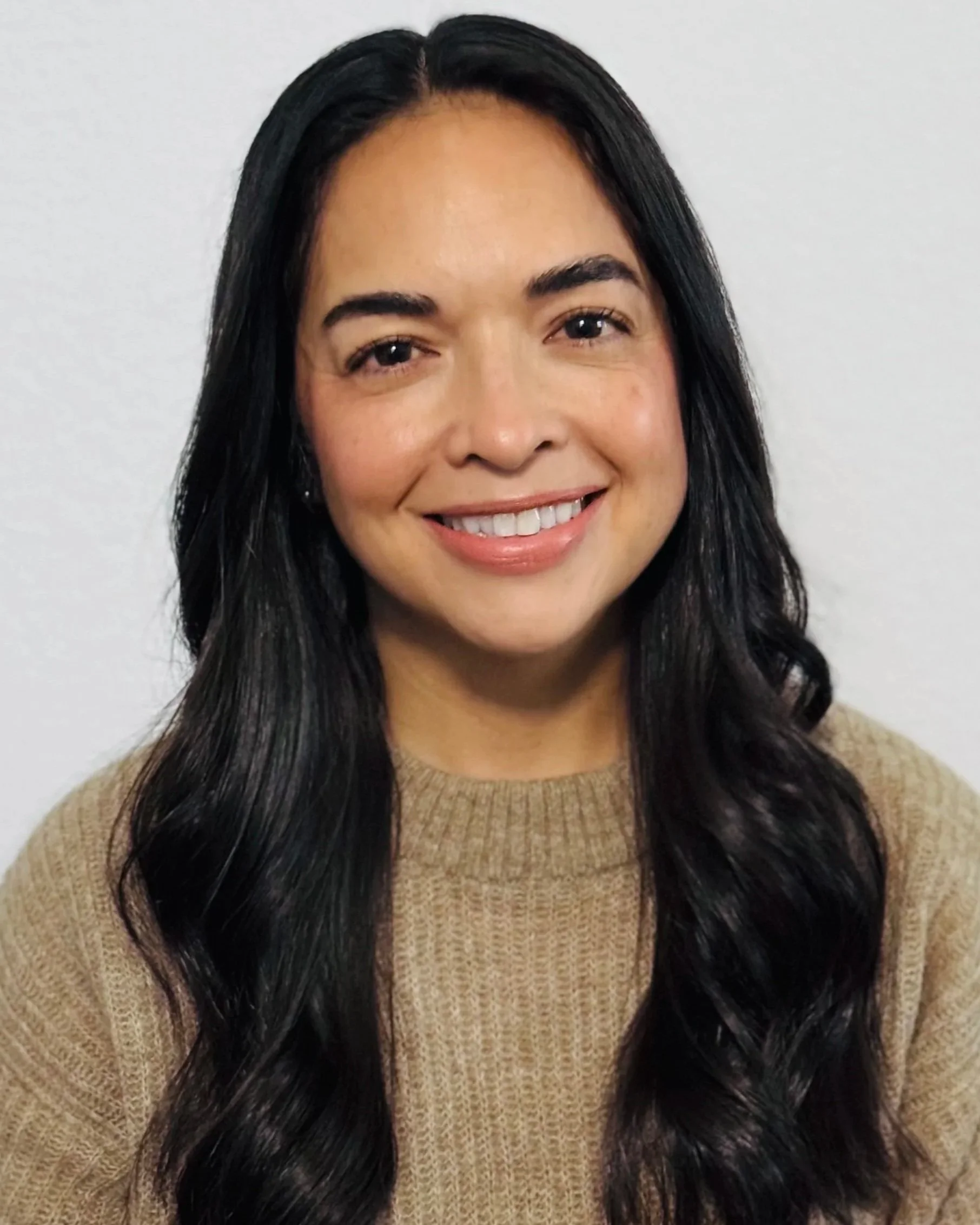 A woman with long black hair, smiling, wearing a beige sweater, standing against a white wall.