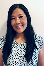 Portrait of a woman with long black hair smiling, wearing a white blouse with black polka dots, against a plain white background.