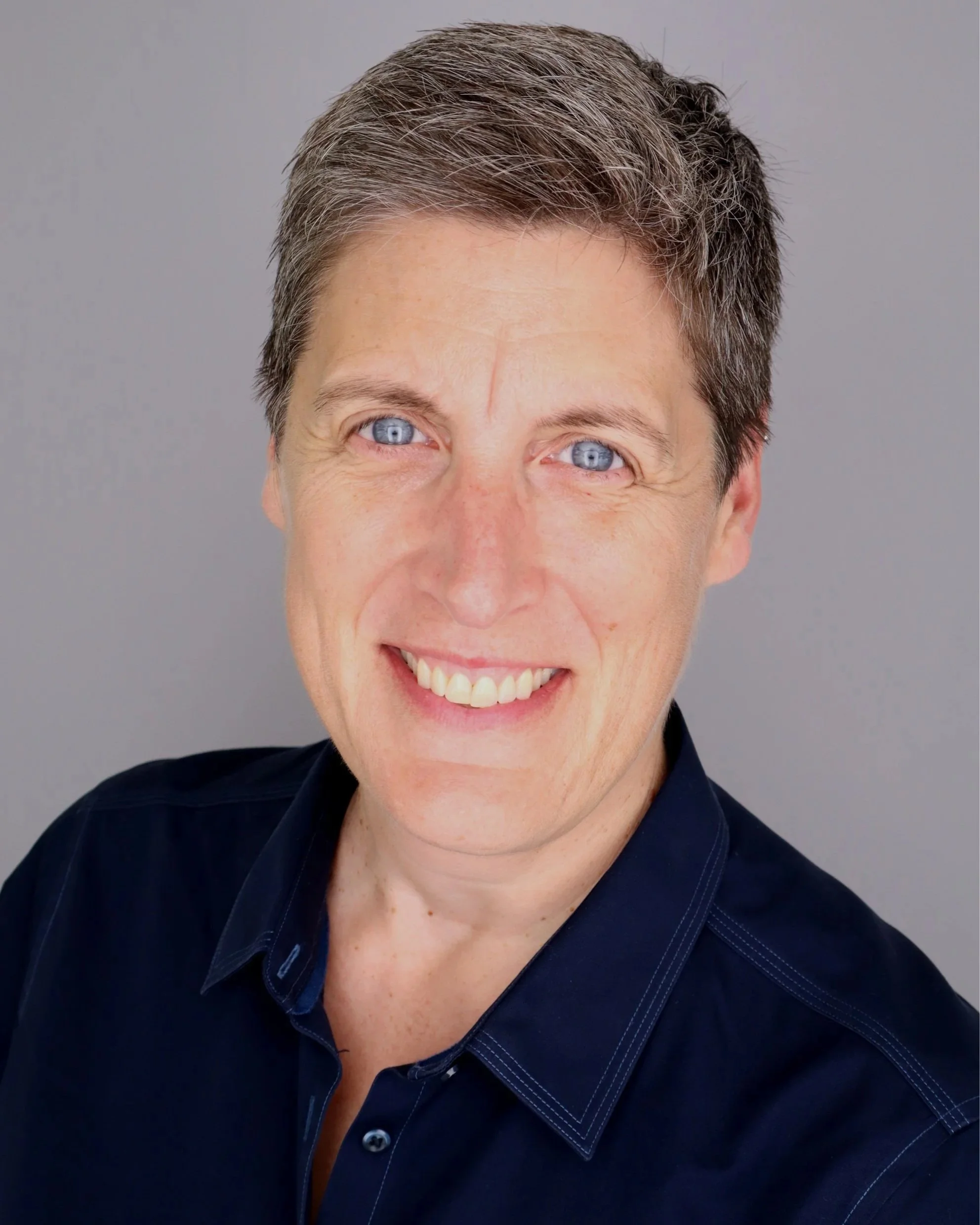A man with short brown hair, blue eyes, and a bright smile, wearing a dark blue collared shirt, standing against a gray background.