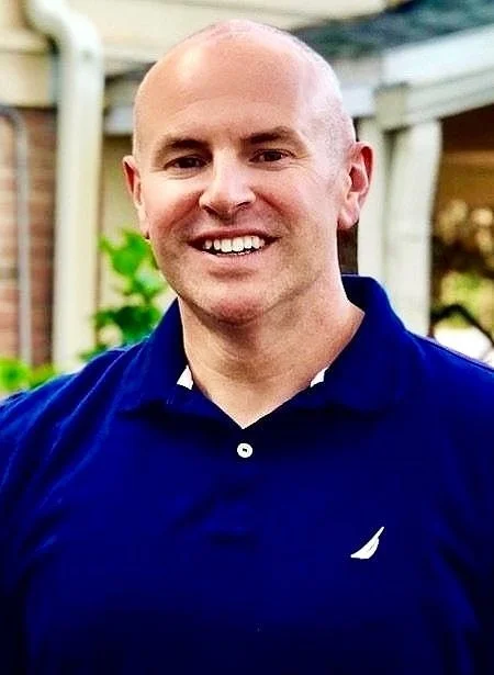 A smiling man with a bald head wearing a navy blue polo shirt outdoors, with a blurred background of trees and a house.