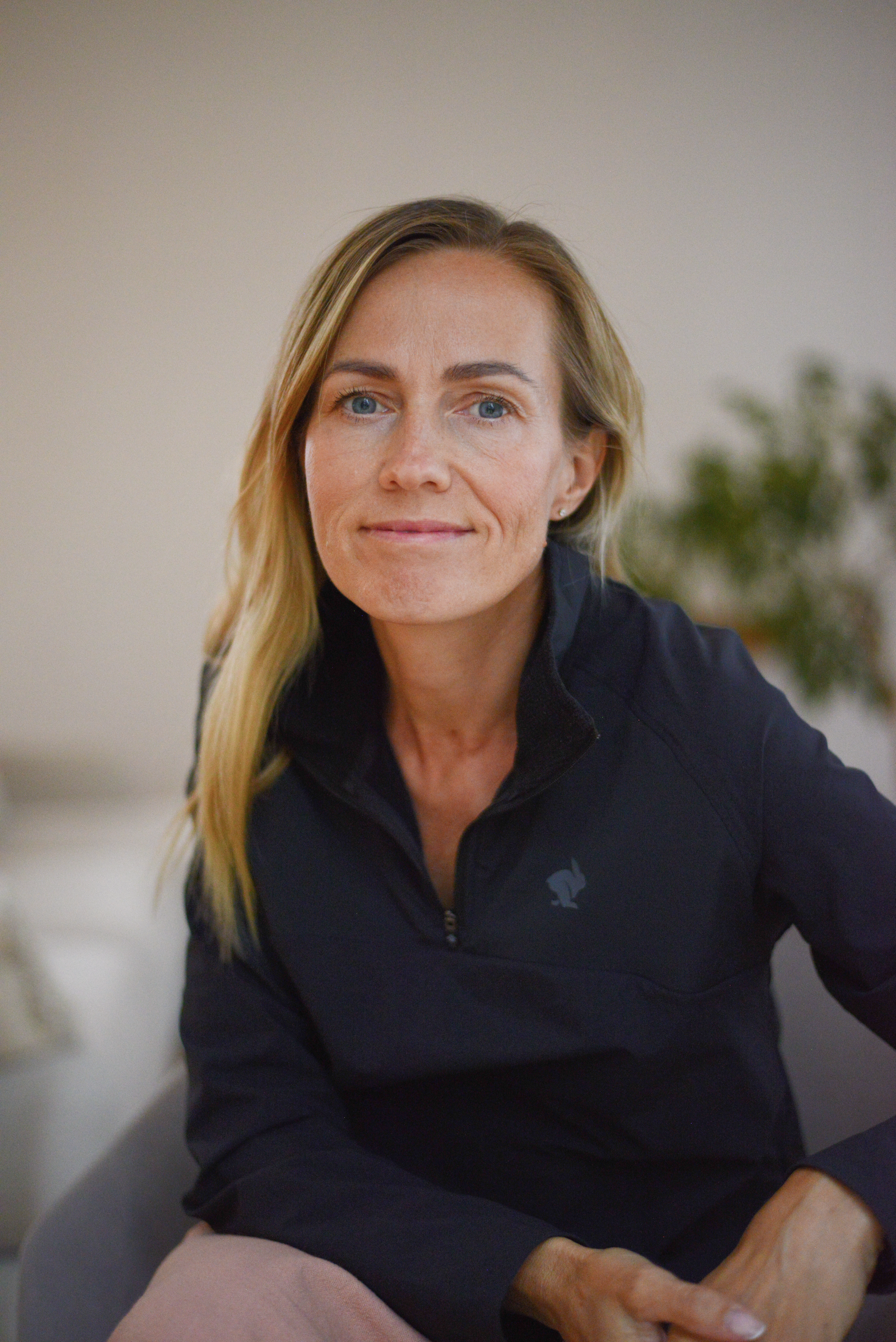 A woman with long blonde hair, blue eyes, and fair skin sits indoors. She is wearing a black zip-up jacket and appears to be smiling softly. In the background, there is a blurred plant and neutral-colored wall.
