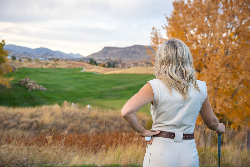 A woman with blonde, wavy hair standing on a golf course, looking toward the distance with an autumn landscape in the background.