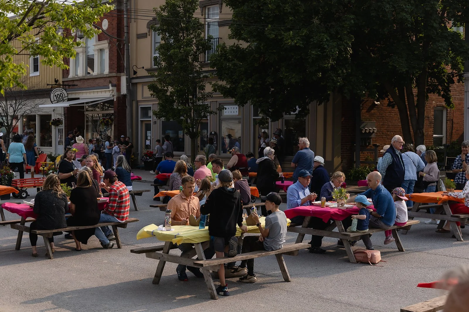 Outdoor community event with people sitting at picnic tables on a street, surrounded by buildings and trees. Participants are socializing and enjoying food in creemore. Town and county collective media agency