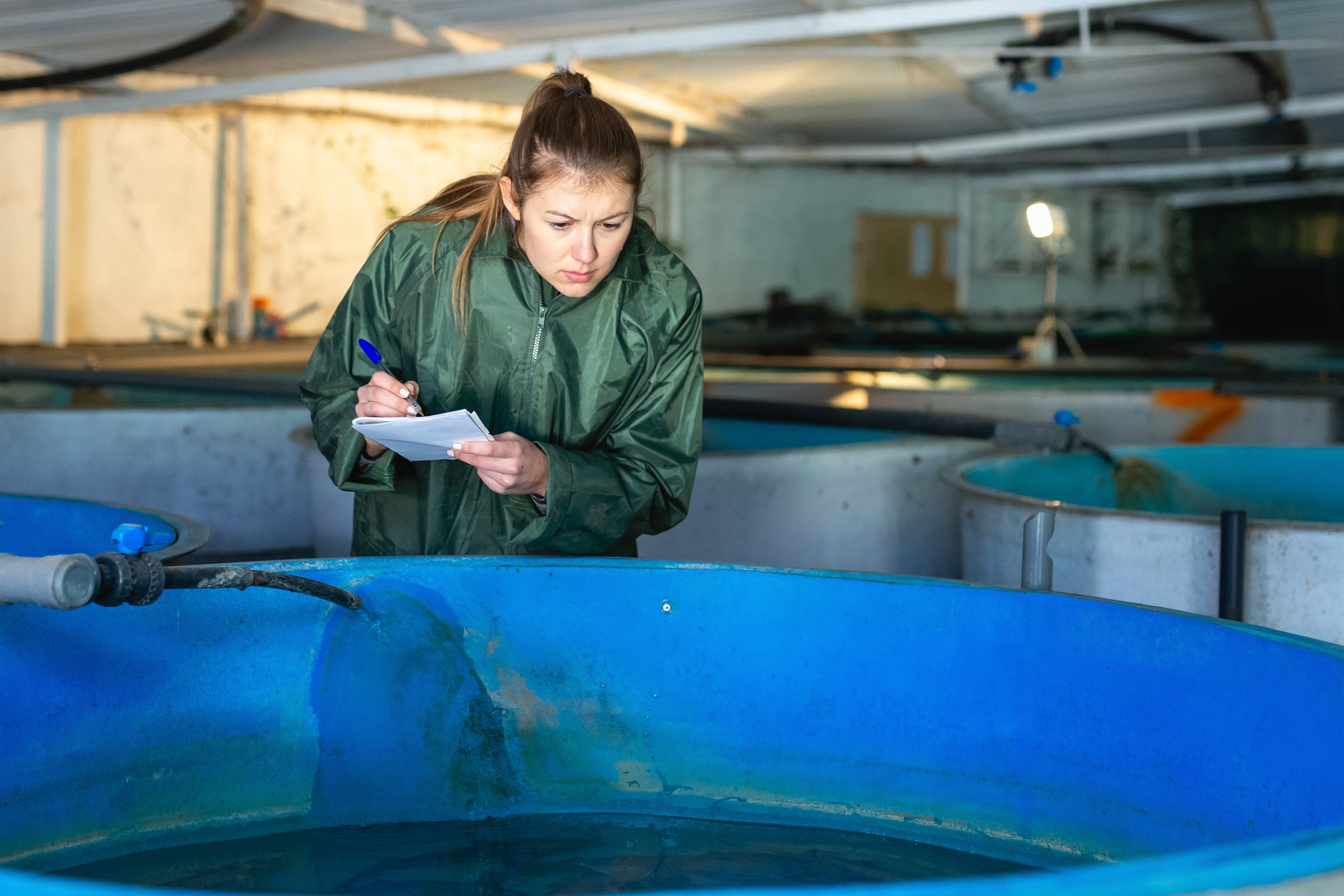 A woman in a green jacket is inspecting or taking notes at circular blue tanks in an indoor aquaculture or fish farming facility.