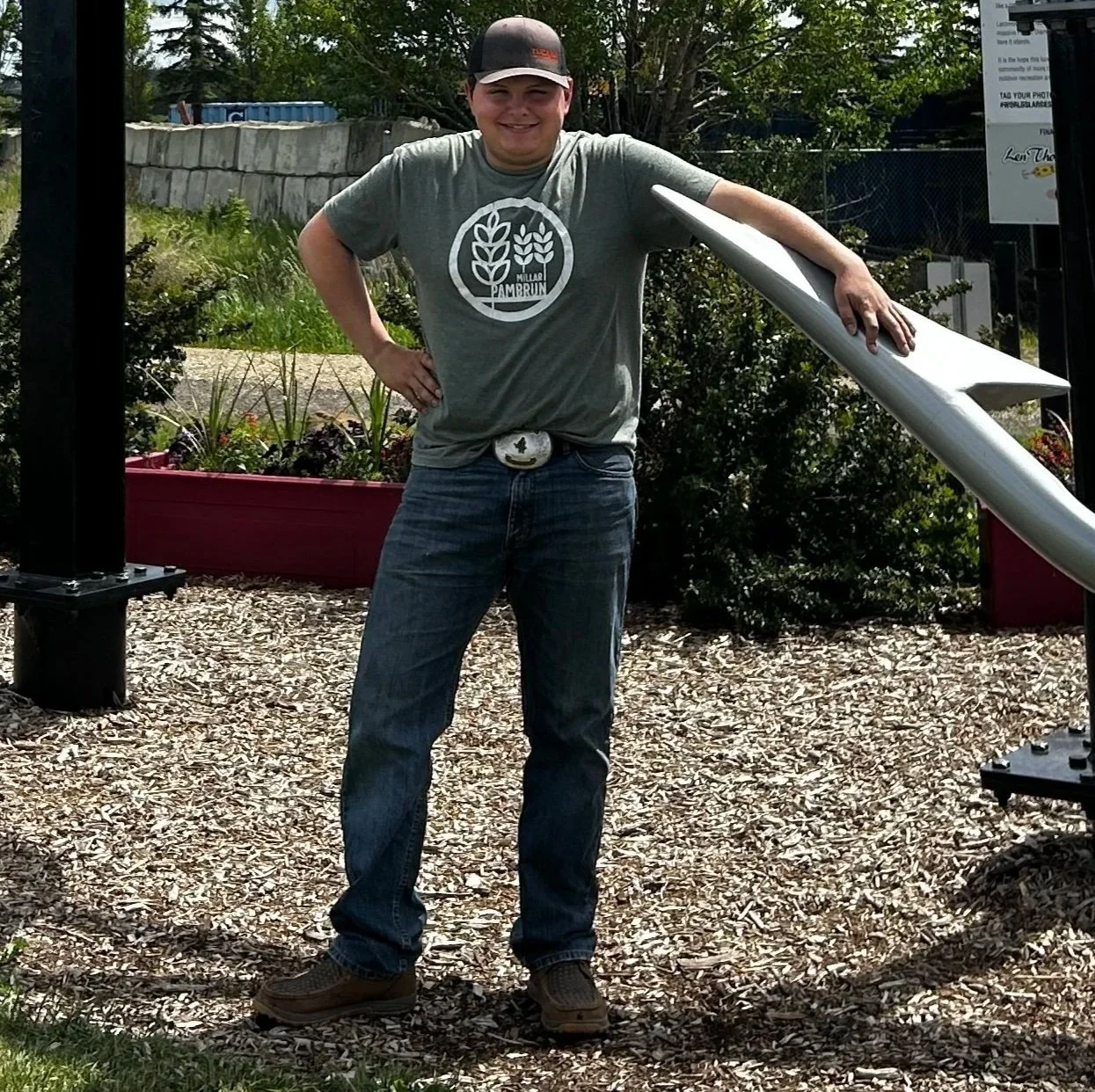 A smiling man wearing a gray cap and sunglasses hanging from his shirt, giving two thumbs up in a field with green plants and a forest of trees in the background under a clear blue sky.
