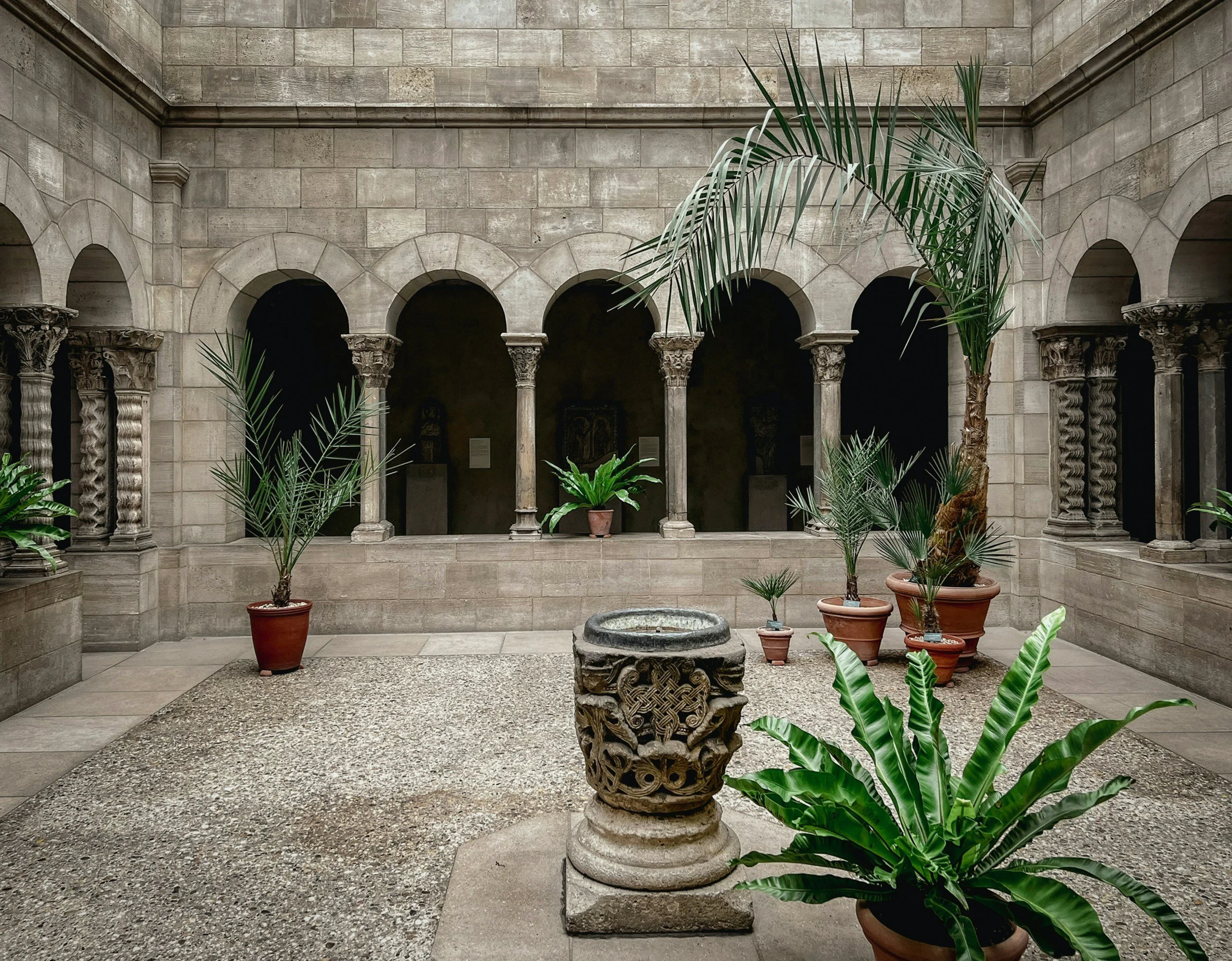 Interior of a historic stone building with arched windows and decorative columns, featuring potted plants and a carved stone pedestal in the center.