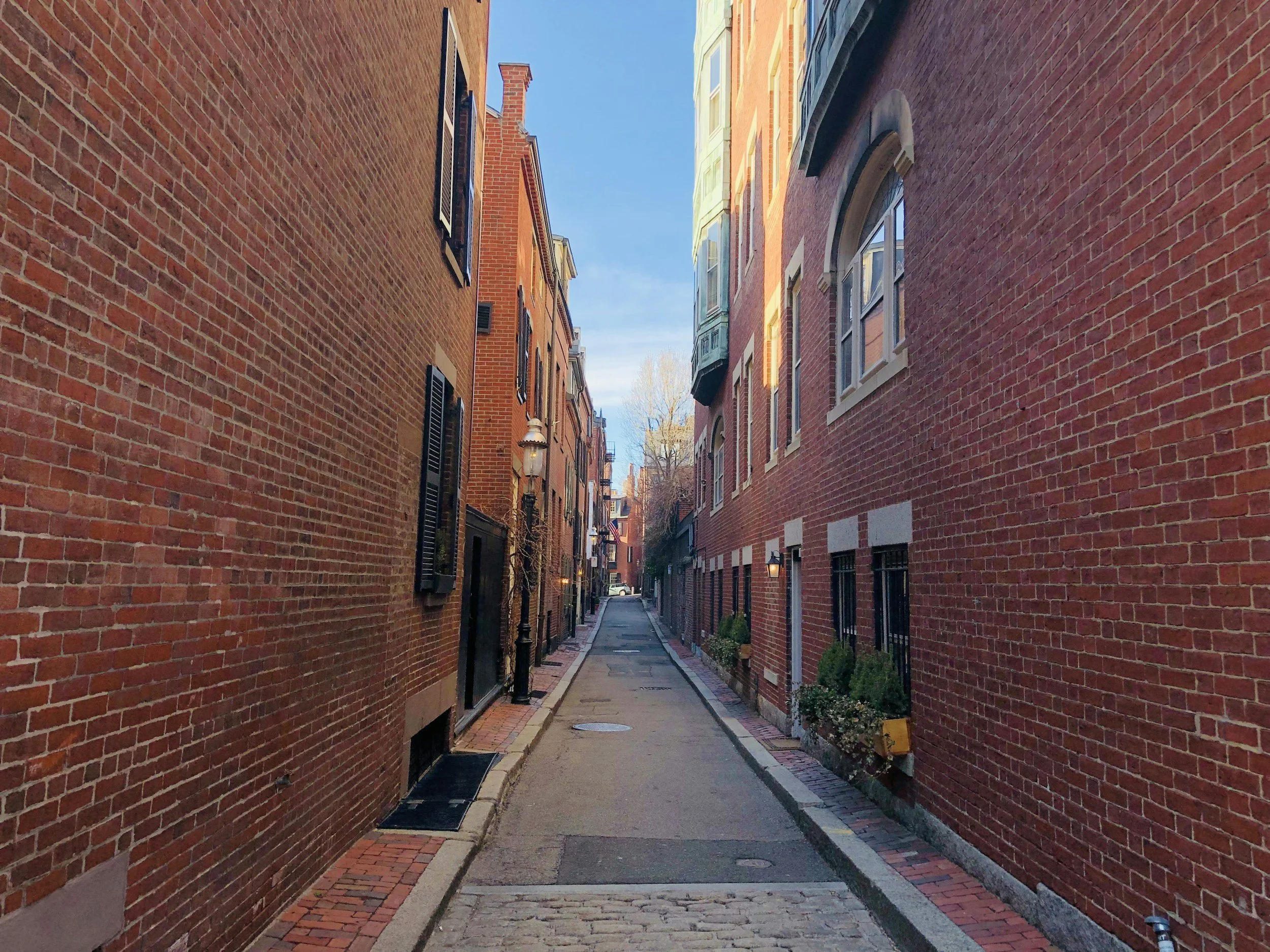 A narrow alleyway between brick buildings with black shutters and street lamps, leading to a small tree and colorful buildings in the distance.