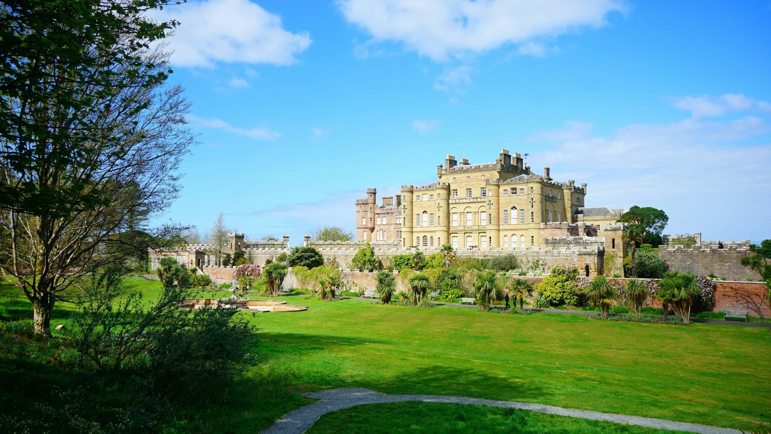 A large historic castle surrounded by lush green gardens, with a bright blue sky and some clouds overhead.