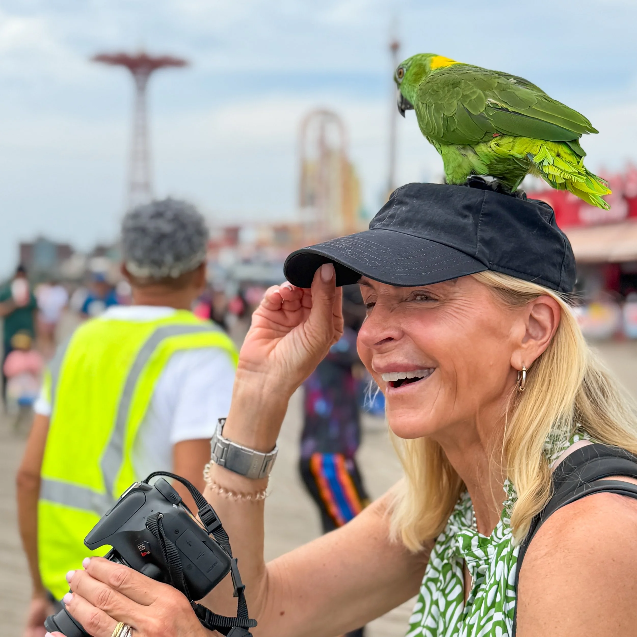 A woman in a green and white patterned dress is adjusting a black hat with her right hand while a yellow-green parrot is perched on her left side. The woman is smiling and wearing a watch and a bracelet.