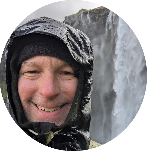 A man smiling and dressed in rain gear with a waterfall in the background.