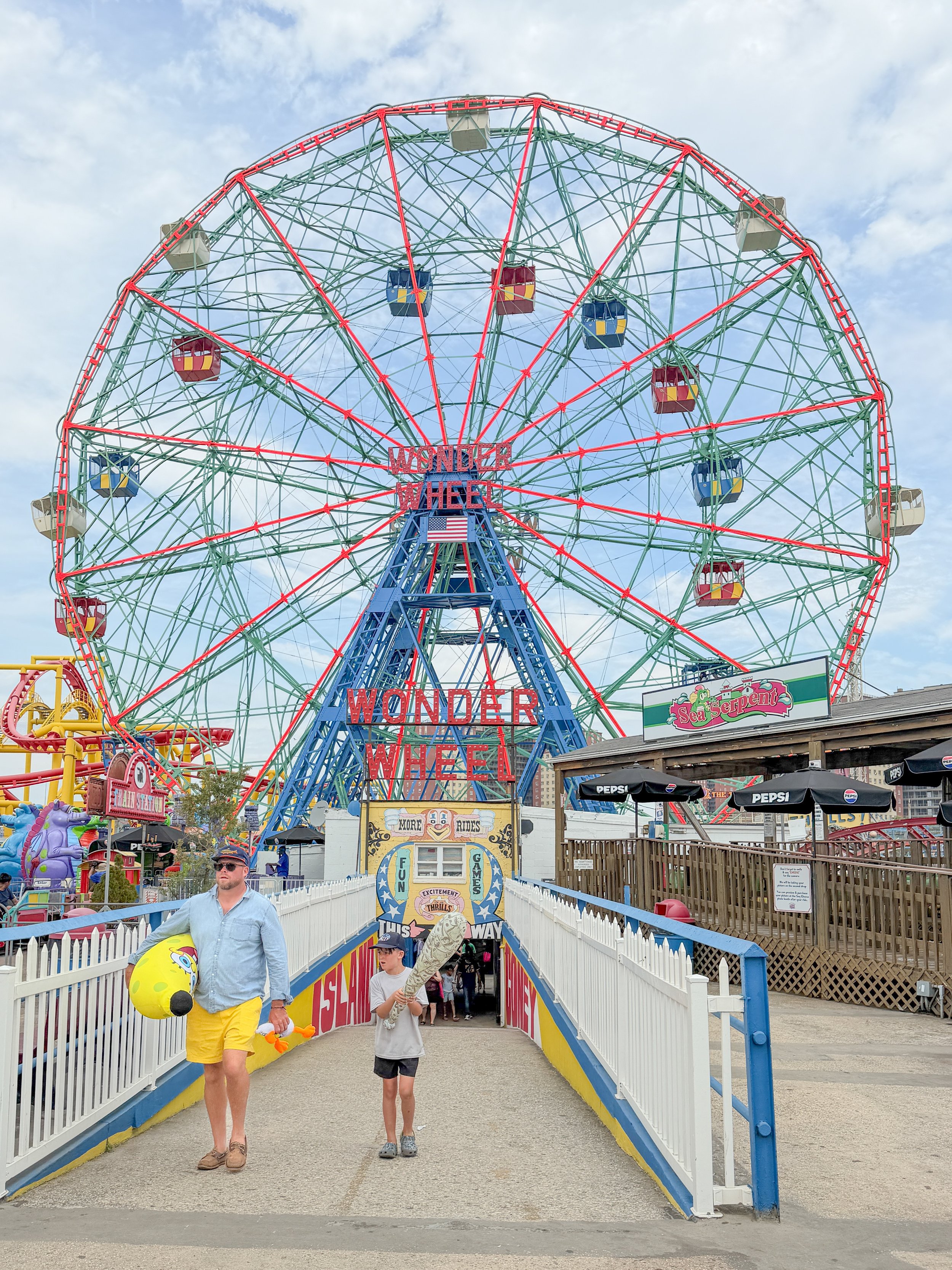 People walking through an entrance to a Ferris wheel at an amusement park called Sea Serpent. The Ferris wheel has colorful gondolas and a sign that reads 'Wonder Wheel'. The sky is partly cloudy, and there are rides and booths visible around the area.