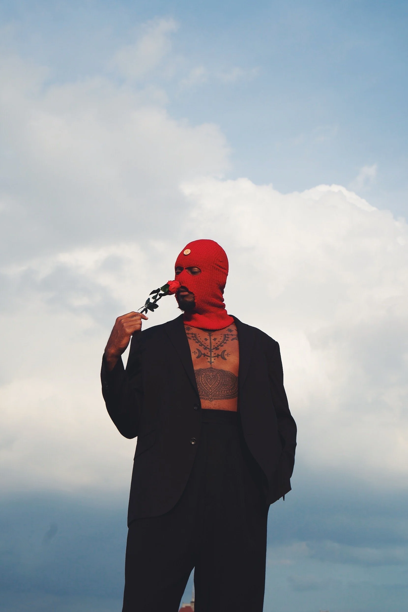 Person wearing a red ski mask, black blazer, and black pants, holding a red rose near their face against a cloudy sky background.