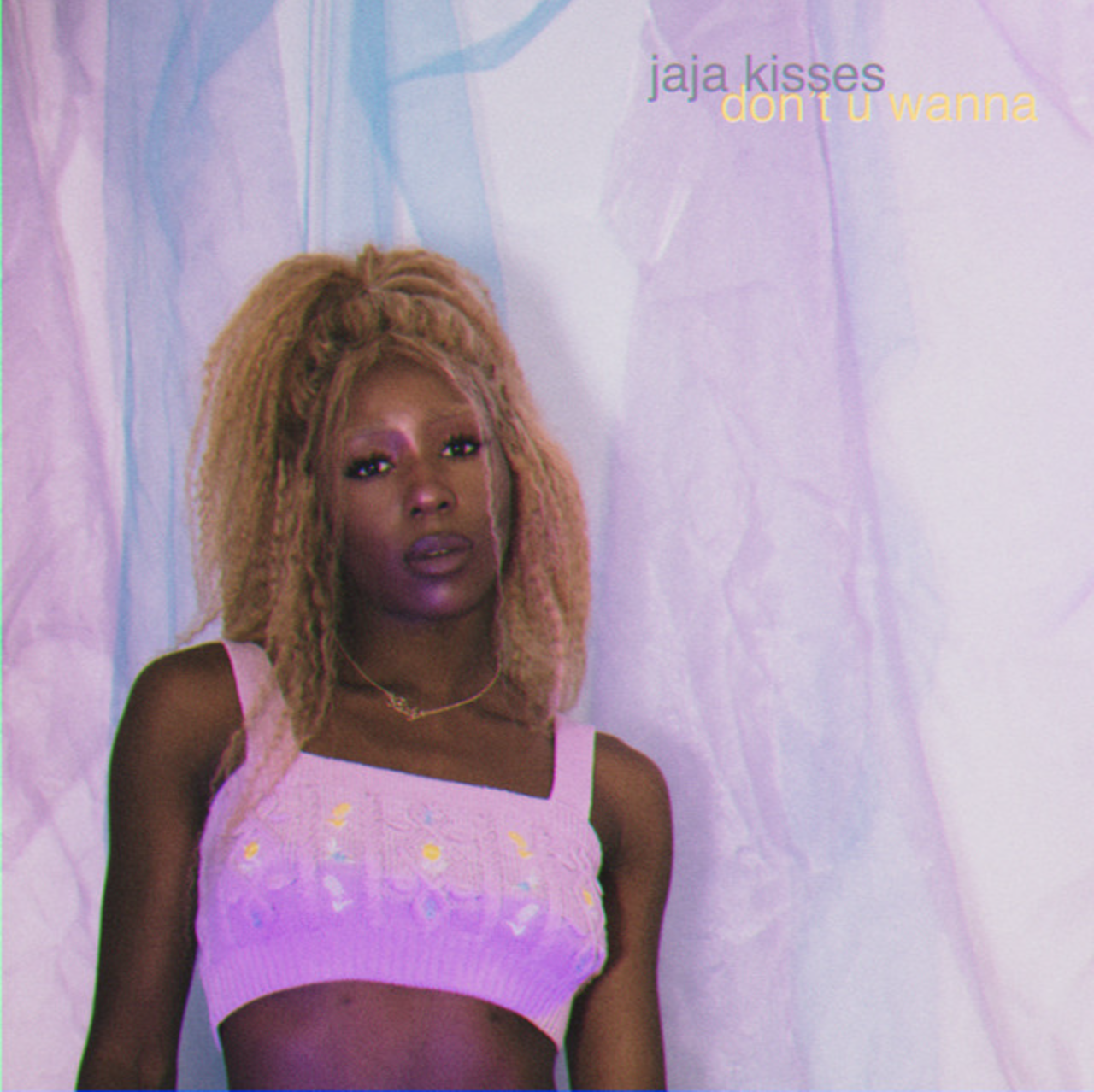 A woman with curly blonde hair wearing a pink embroidered crop top, standing against a pastel-colored background with text in the upper right corner that reads "jaja kisses don't u wanna".