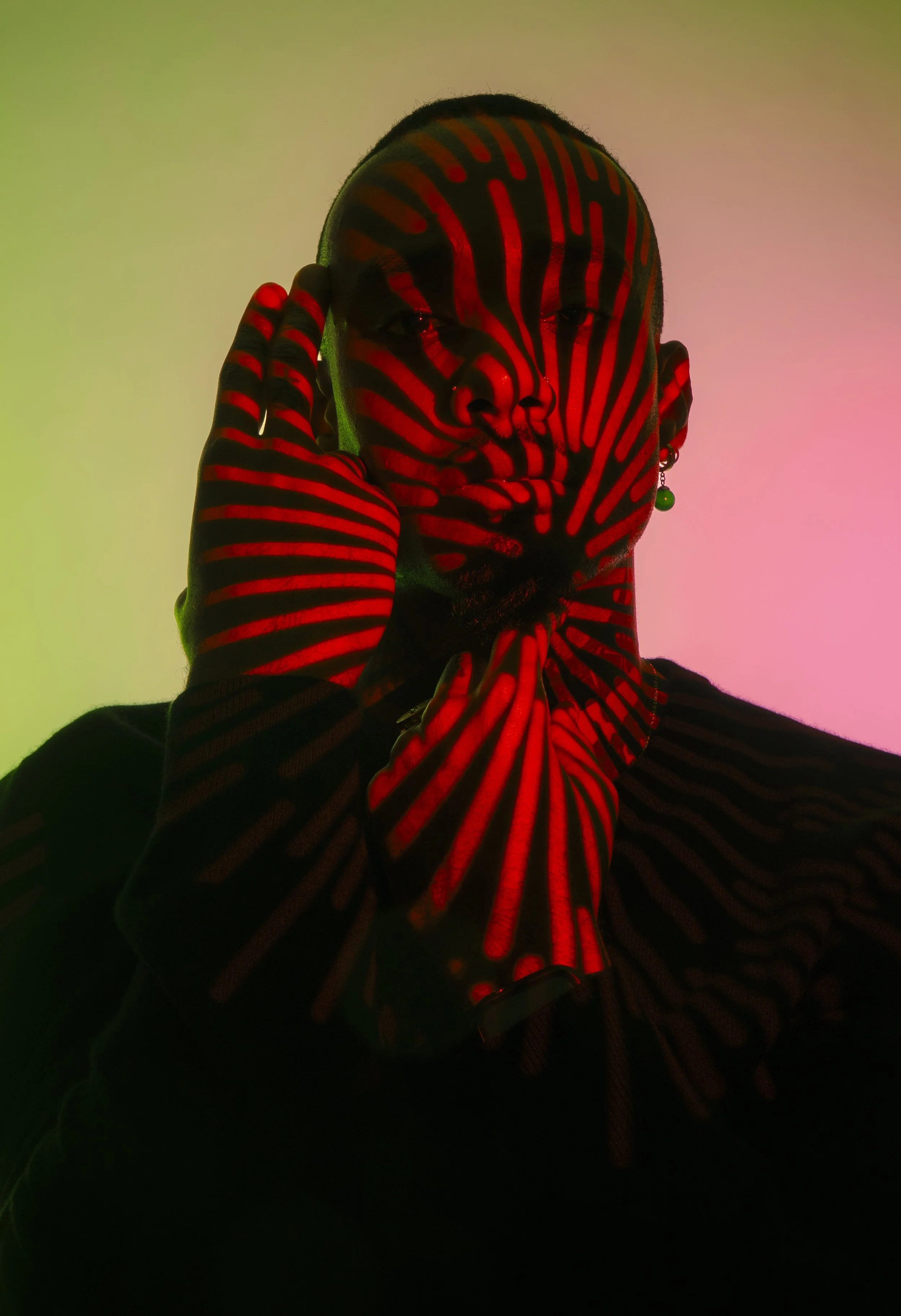 Person with face and hand painted with red and black striped patterns, wearing an earring with a green bead, against a pink and green gradient background.