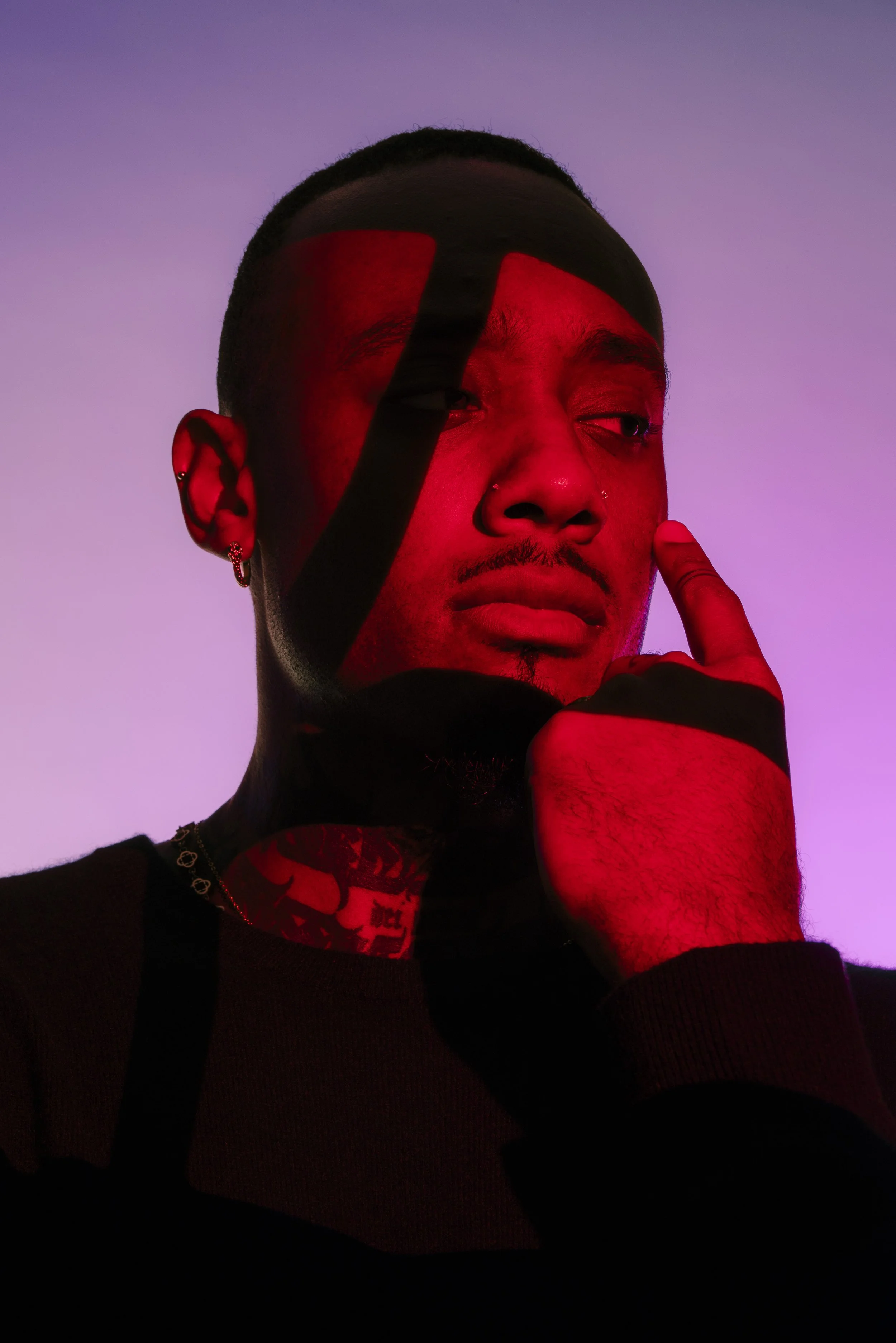 A young man with earrings and tattoos, posing with his finger touching his face against a purple-pink gradient background, partially covered by black and red lighting.