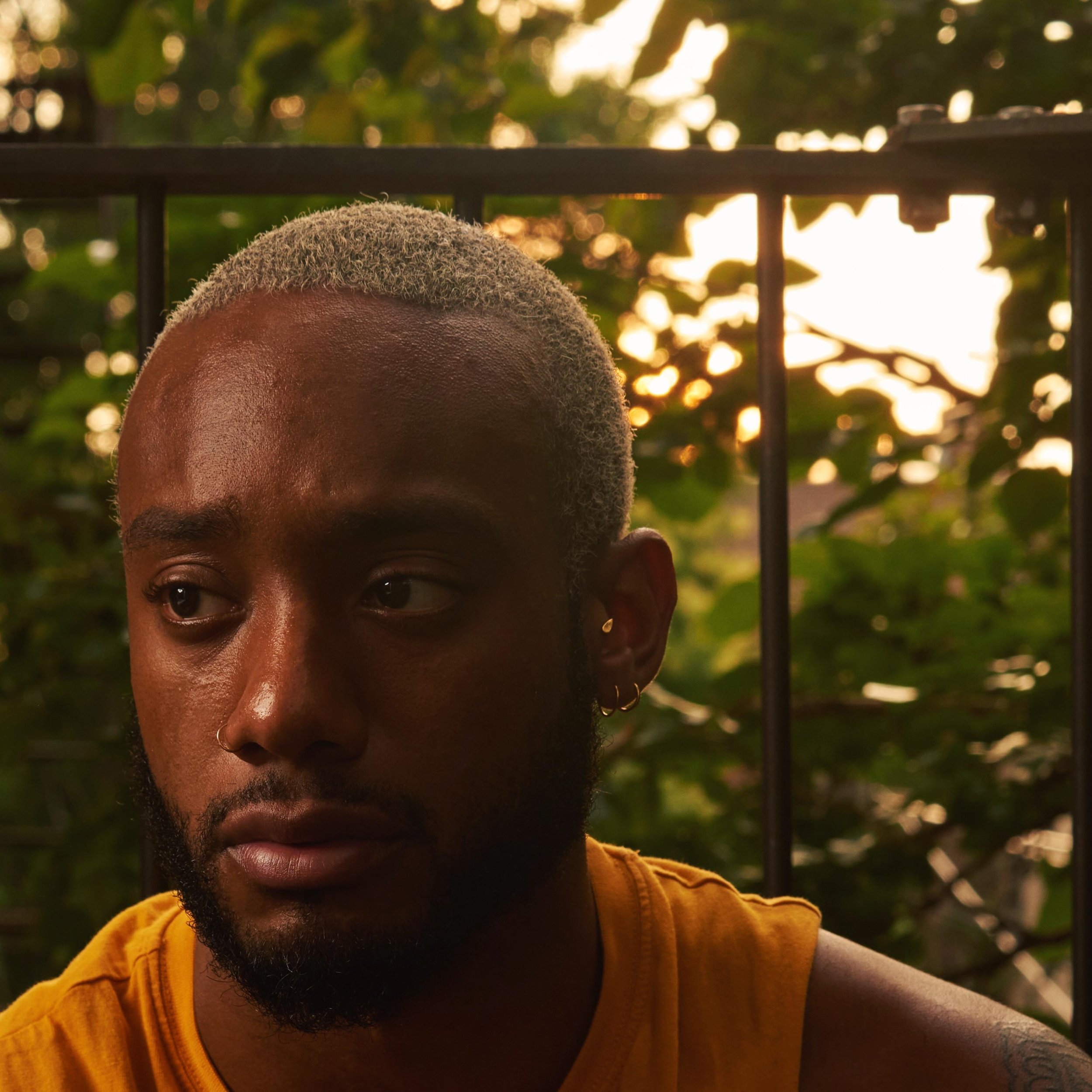 Portrait of a young man with blond hair, beard, and earrings, looking to the side, wearing a yellow shirt, outdoors during sunset with green foliage and metal railing in the background.