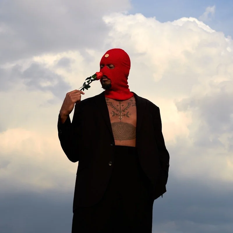 A person wearing a red ski mask and black suit holding a rose close to their face against a cloudy sky background.