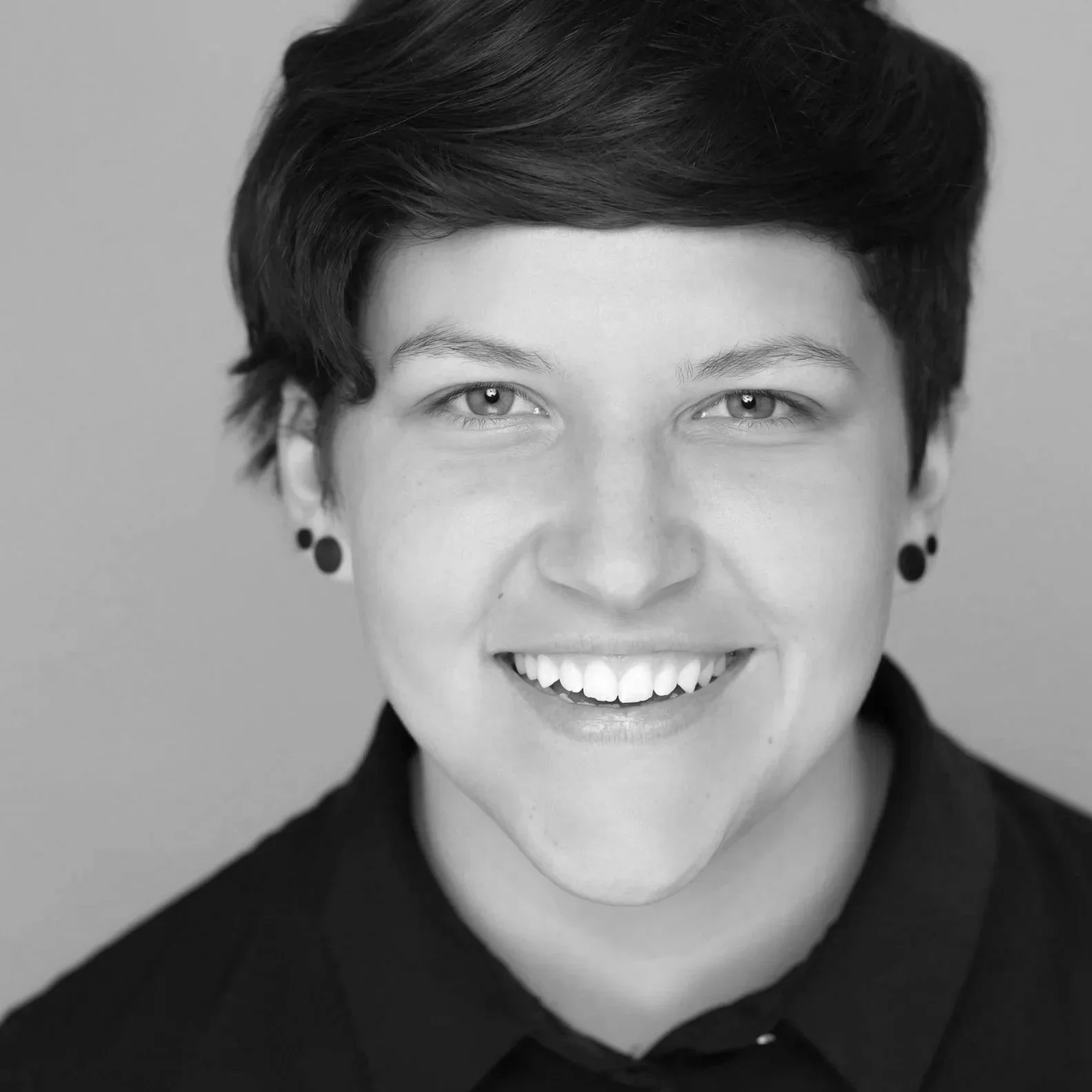 Black and white portrait of a person with short hair, wearing earrings, smiling, and looking directly at the camera.